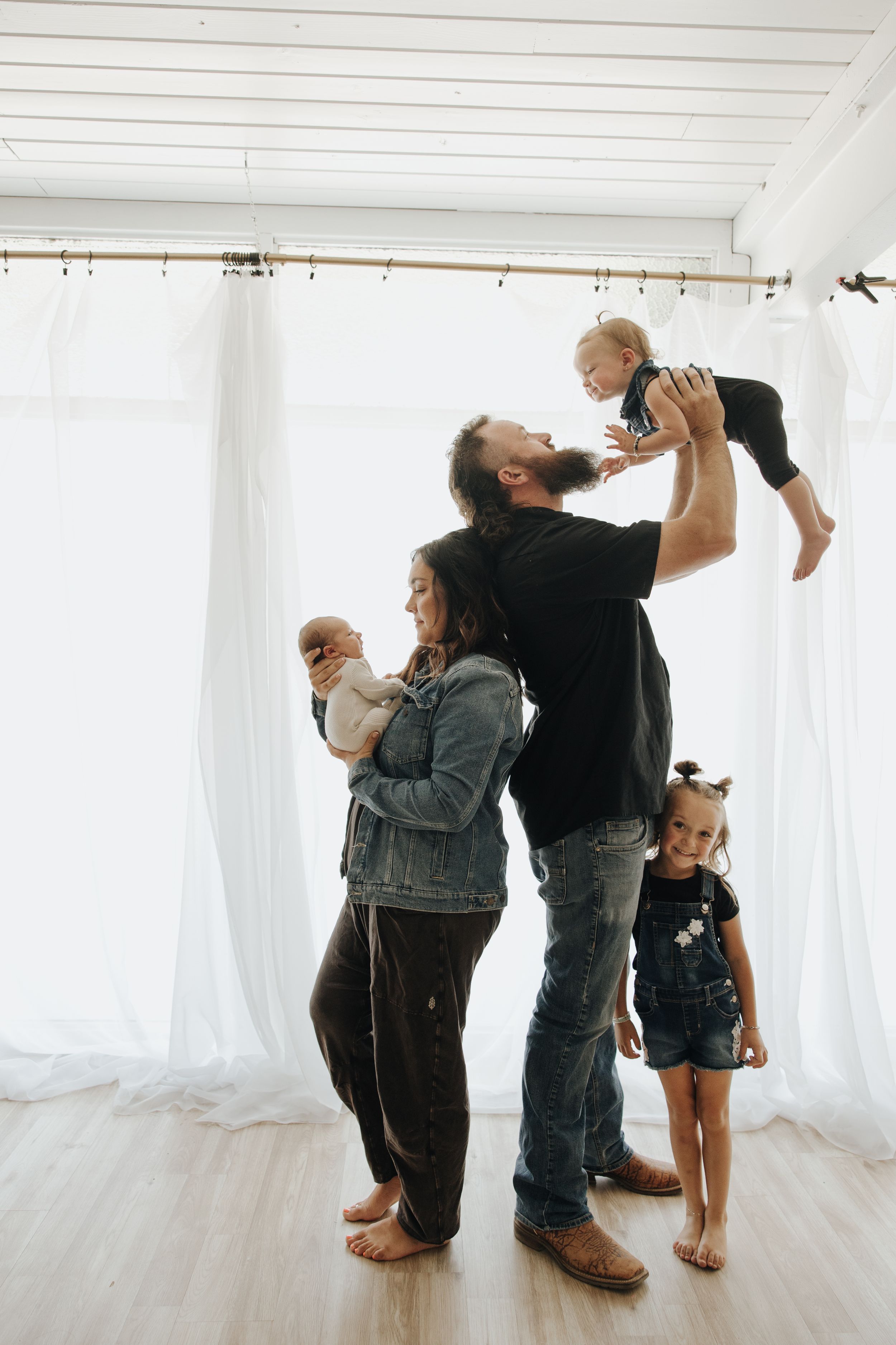 A family lifts and plays with their babies in a bright minimalist room with white walls and wooden floors.