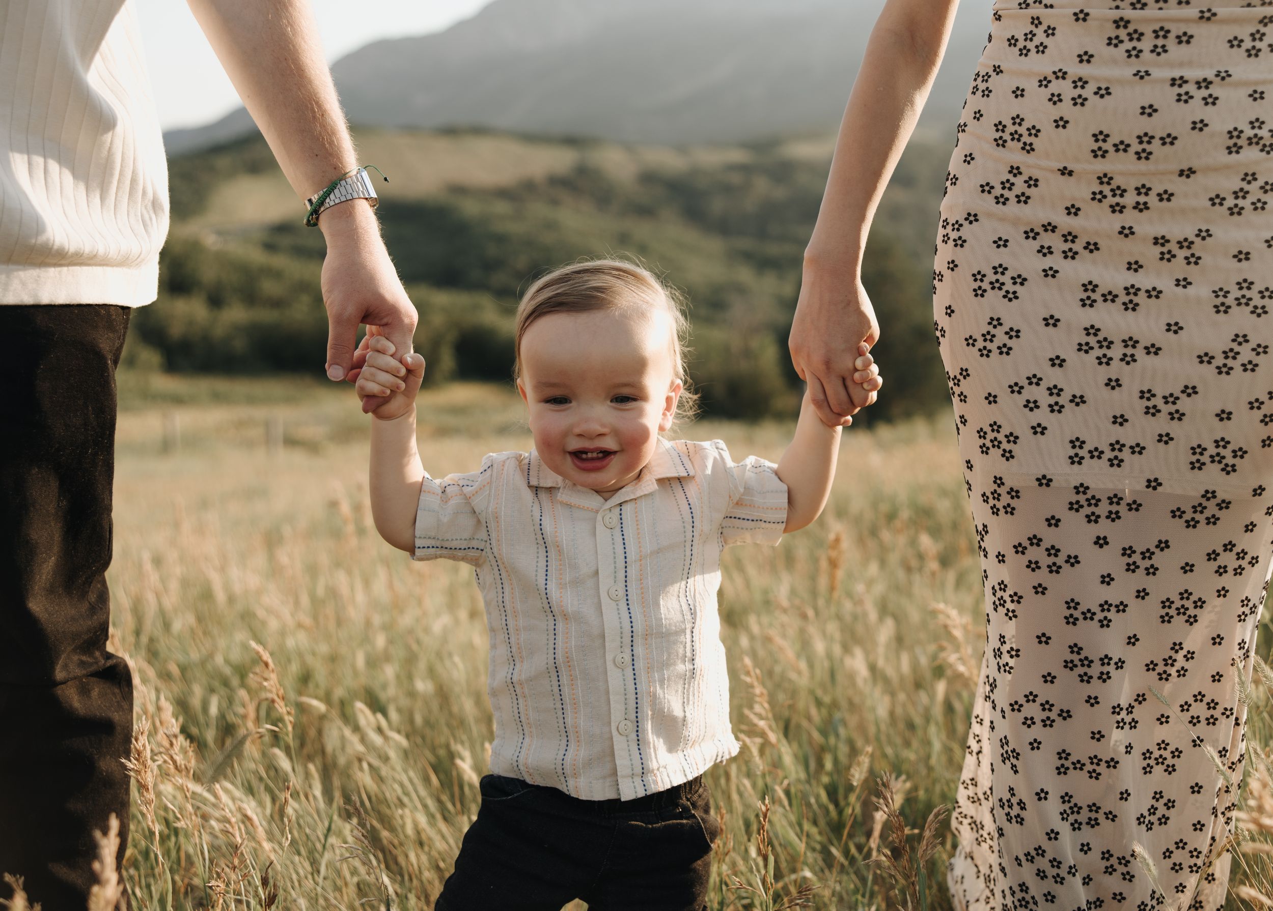 Happy toddler holds hands with parents while walking through a sunlit field.