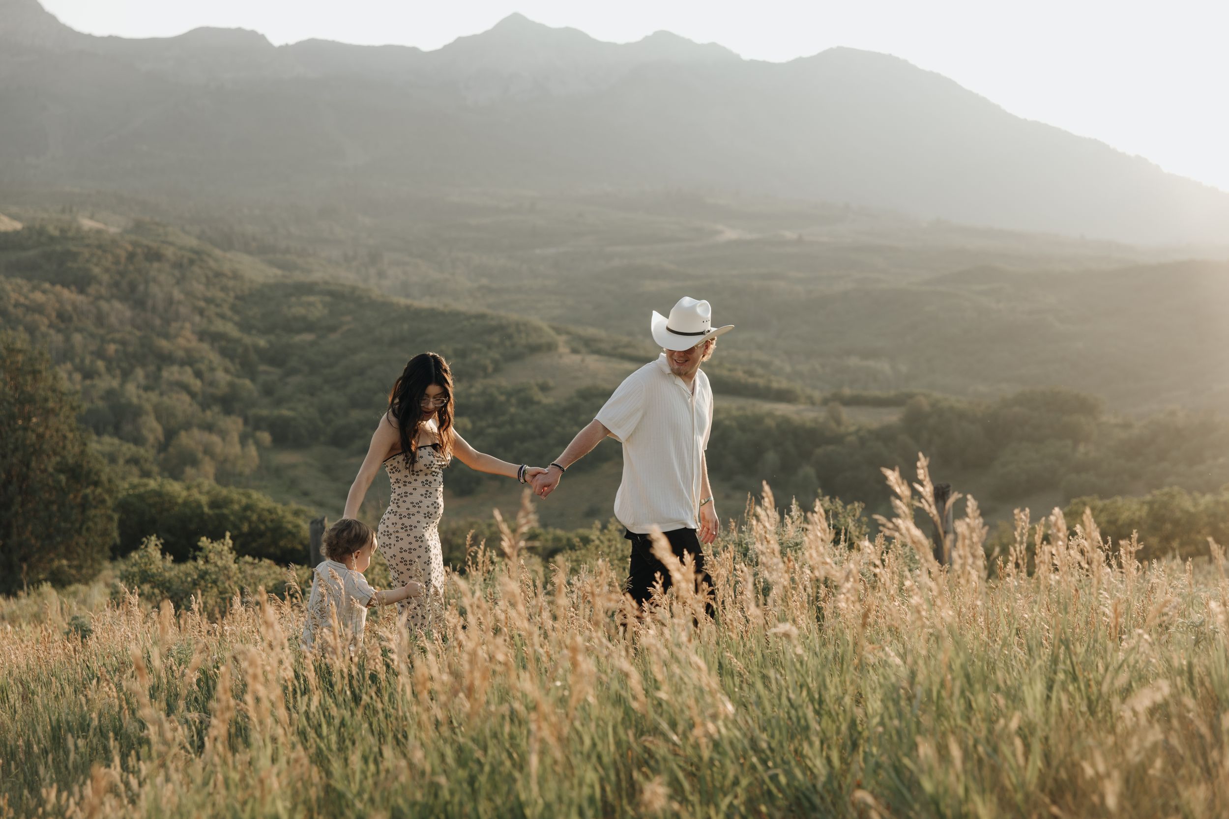 Couple walking hand in hand through tall grass with mountains in the background at sunset.