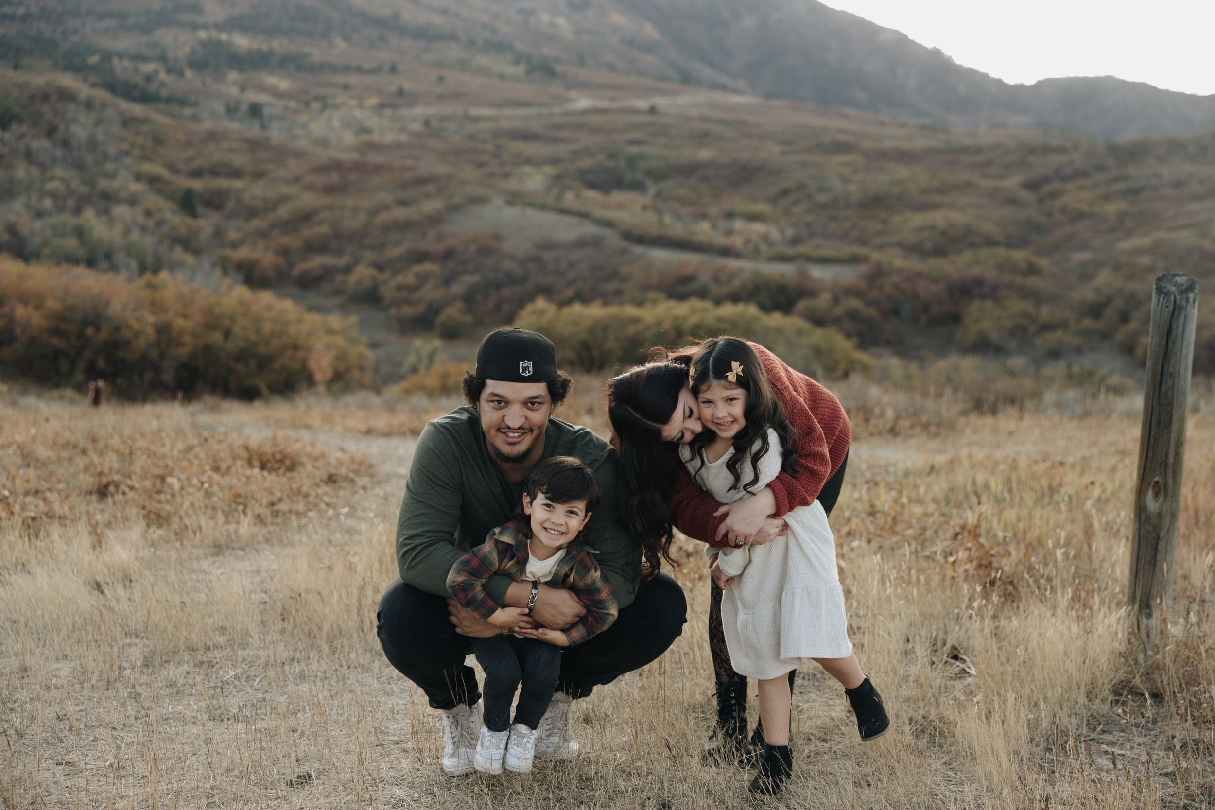 Two people and a husky dog enjoying time together in a golden field with mountains in the background.