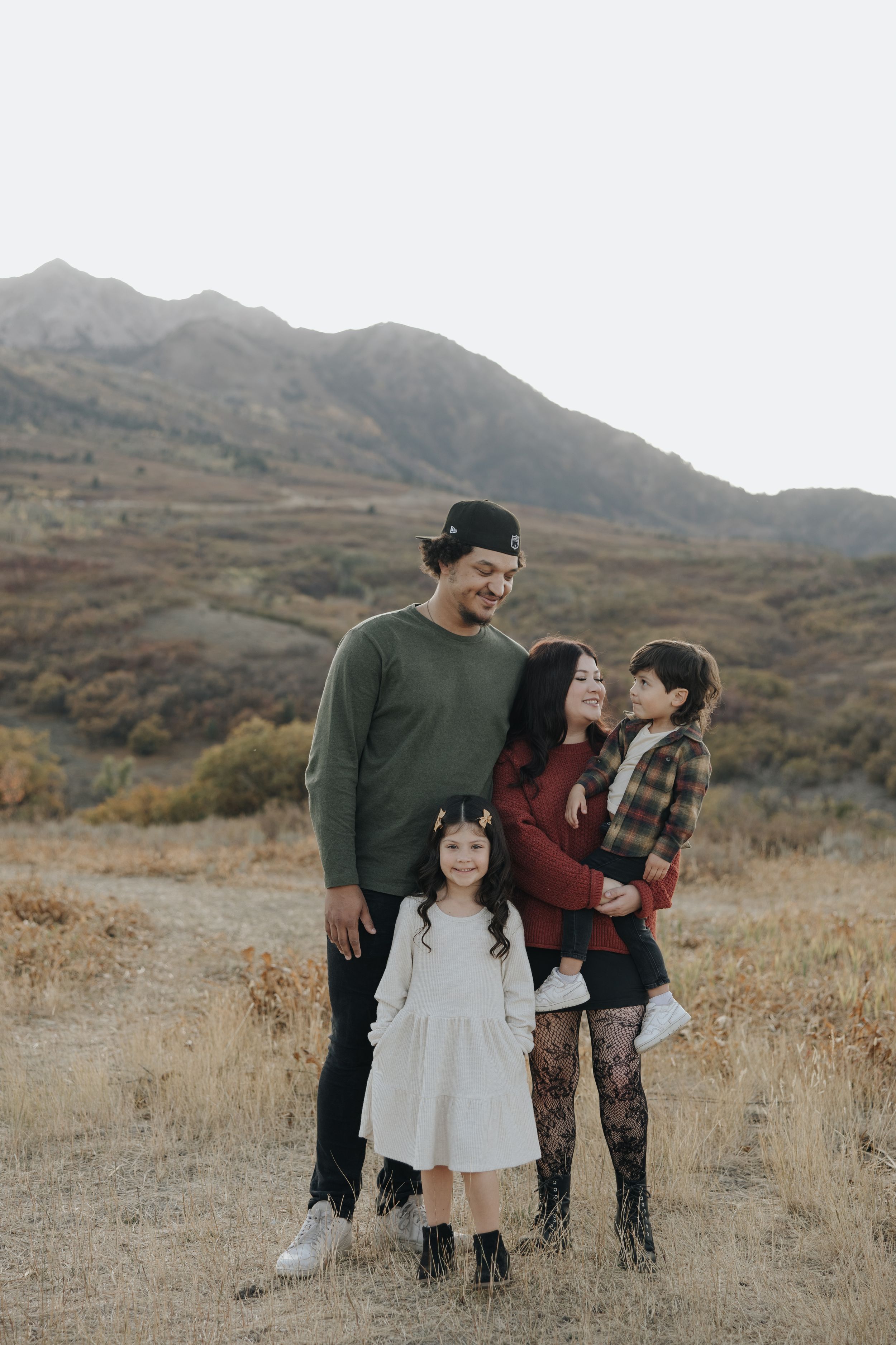 A family stands together in casual fall attire against a backdrop of brown mountains and dry grass.