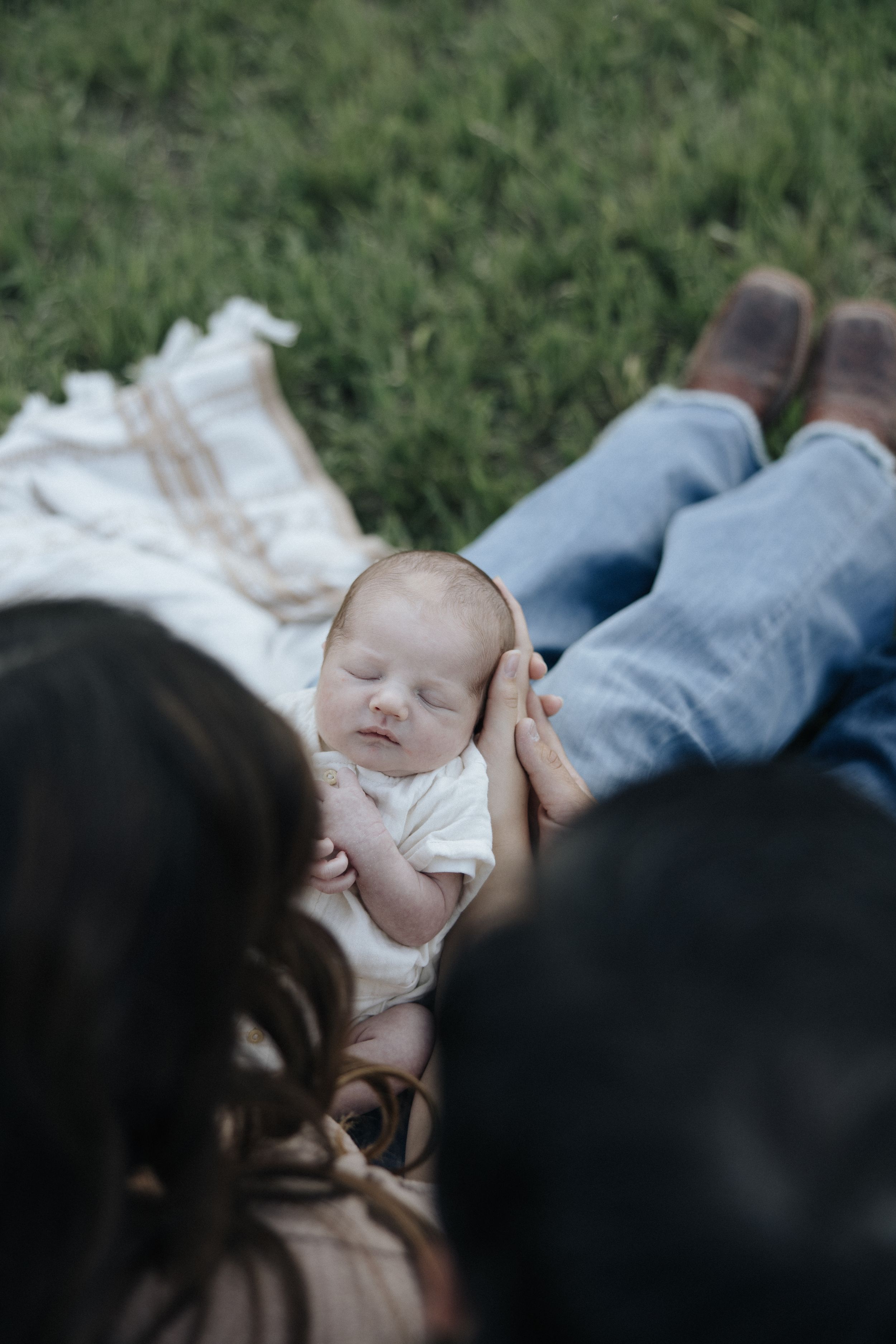 A sleeping baby rests peacefully on a blanket outdoors while two people in jeans look down from above.