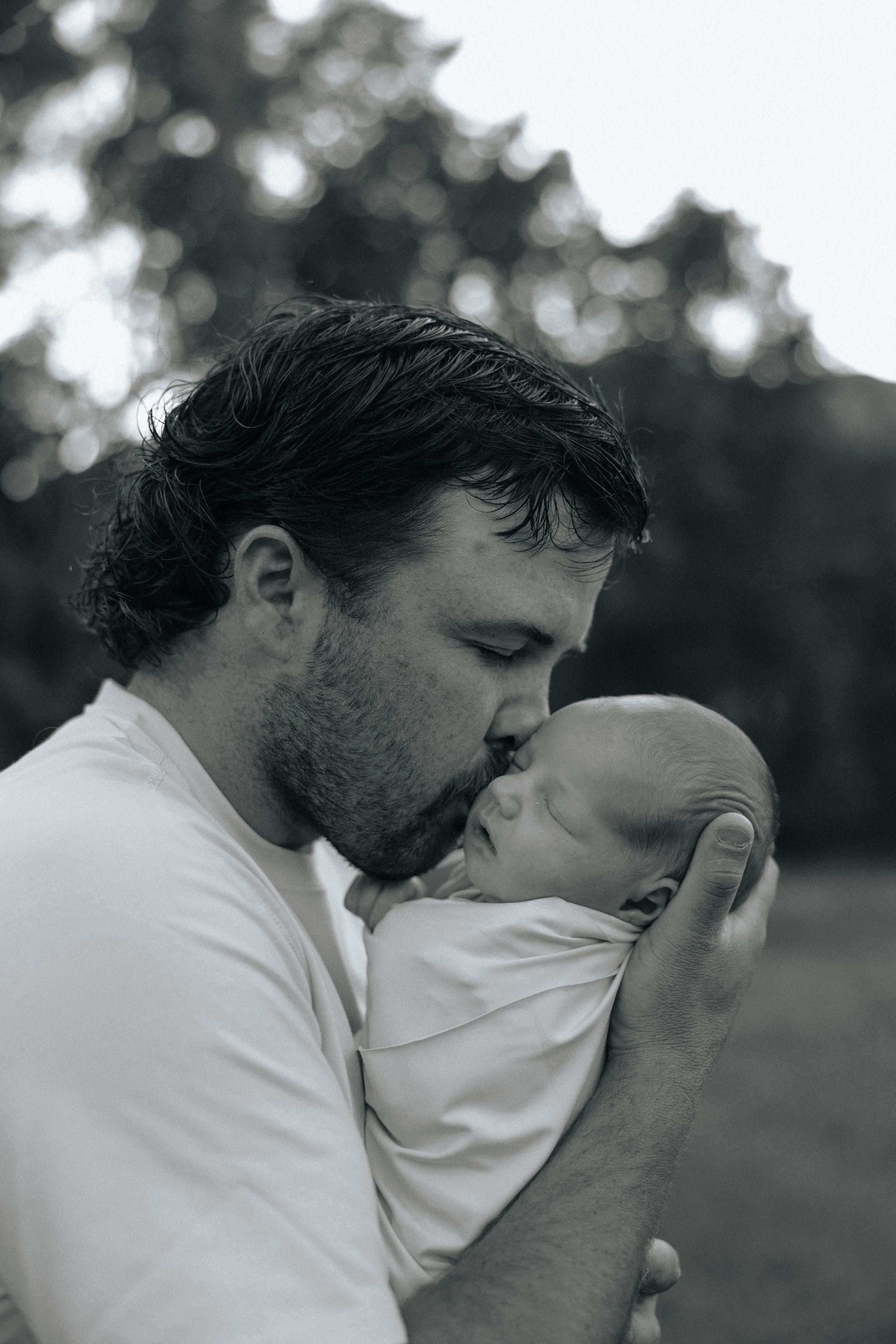 A tender black and white portrait of a father kissing his newborn baby wrapped in a blanket outdoors.