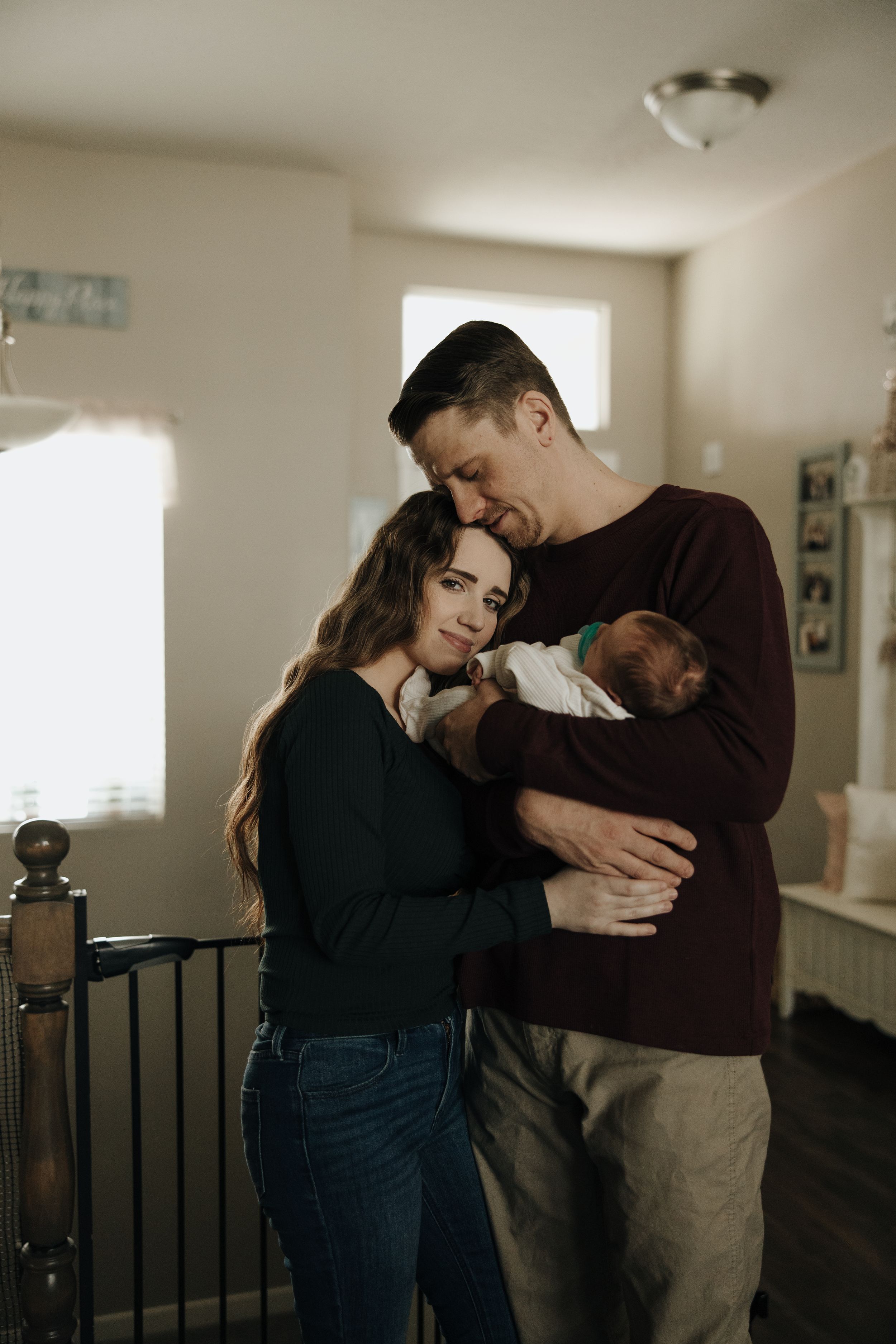 A couple stands together in a cozy room holding their newborn baby in a tender family moment.