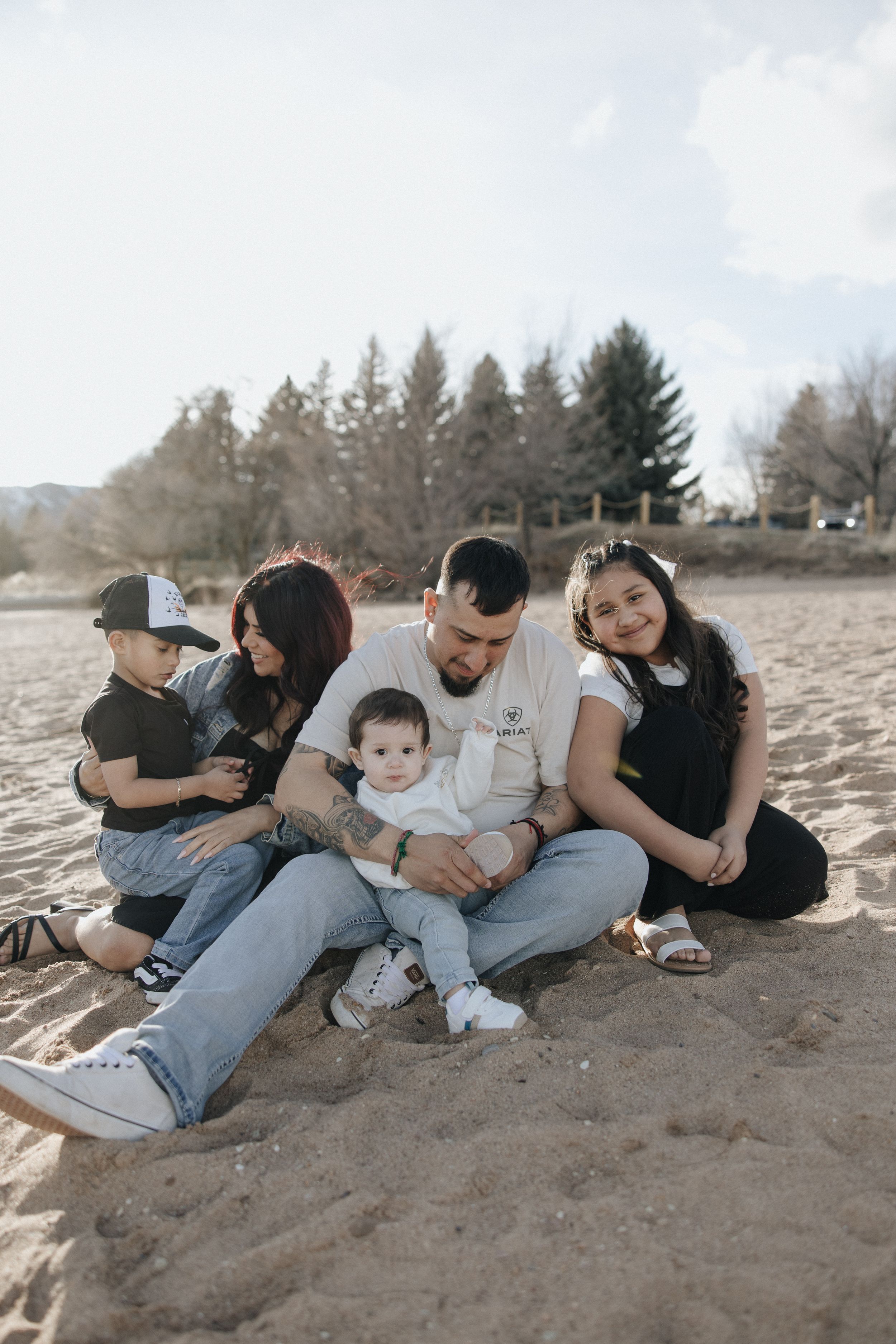 A family sits together on a sandy beach with evergreen trees in the background on a sunny day.
