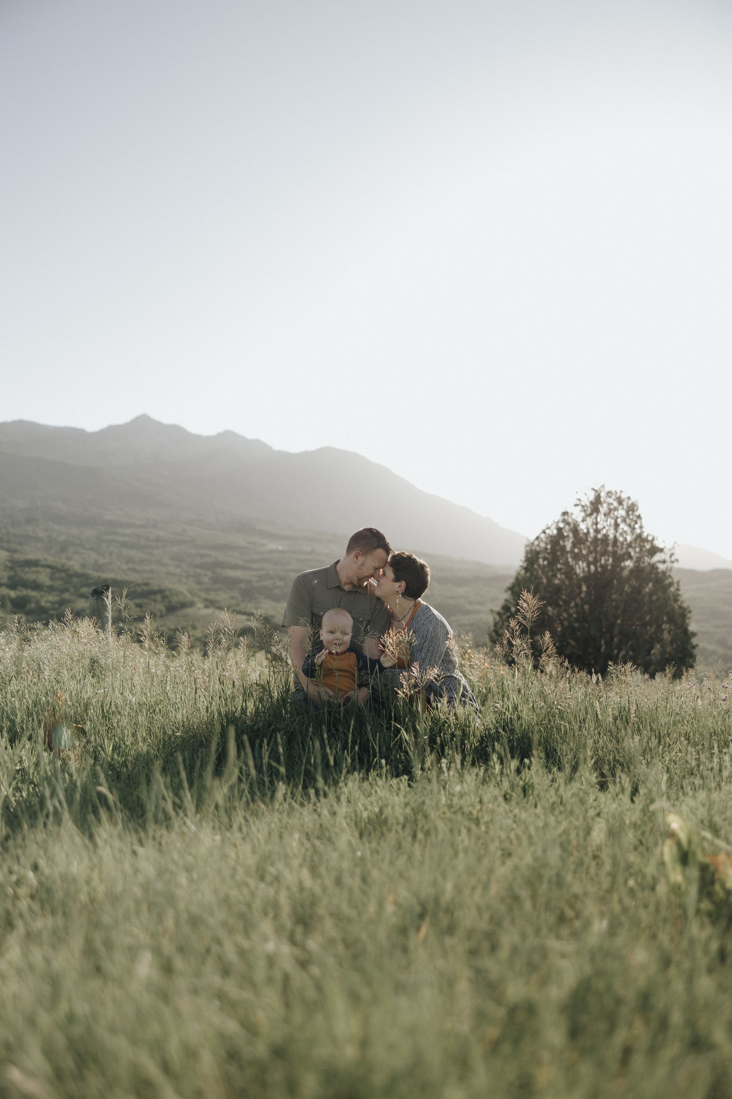 Golden sunlight illuminates a grassy meadow with rolling mountains in the background as figures embrace at sunset.