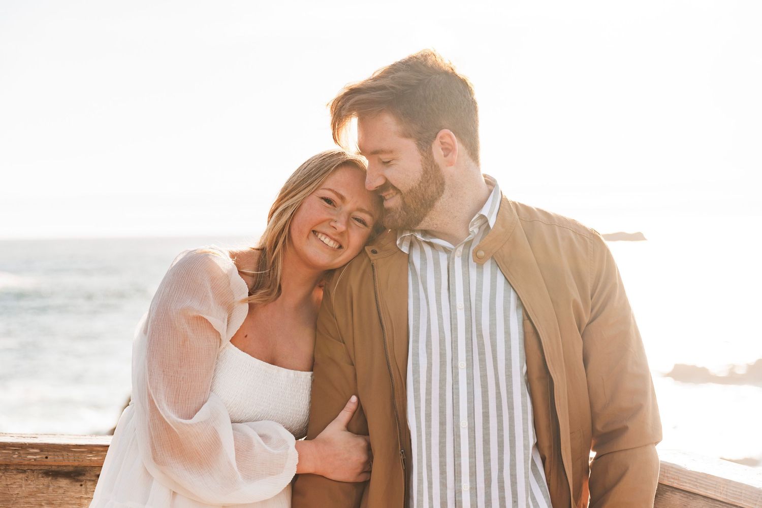 Smiling couple cuddling together by the ocean at golden hour on the coast.