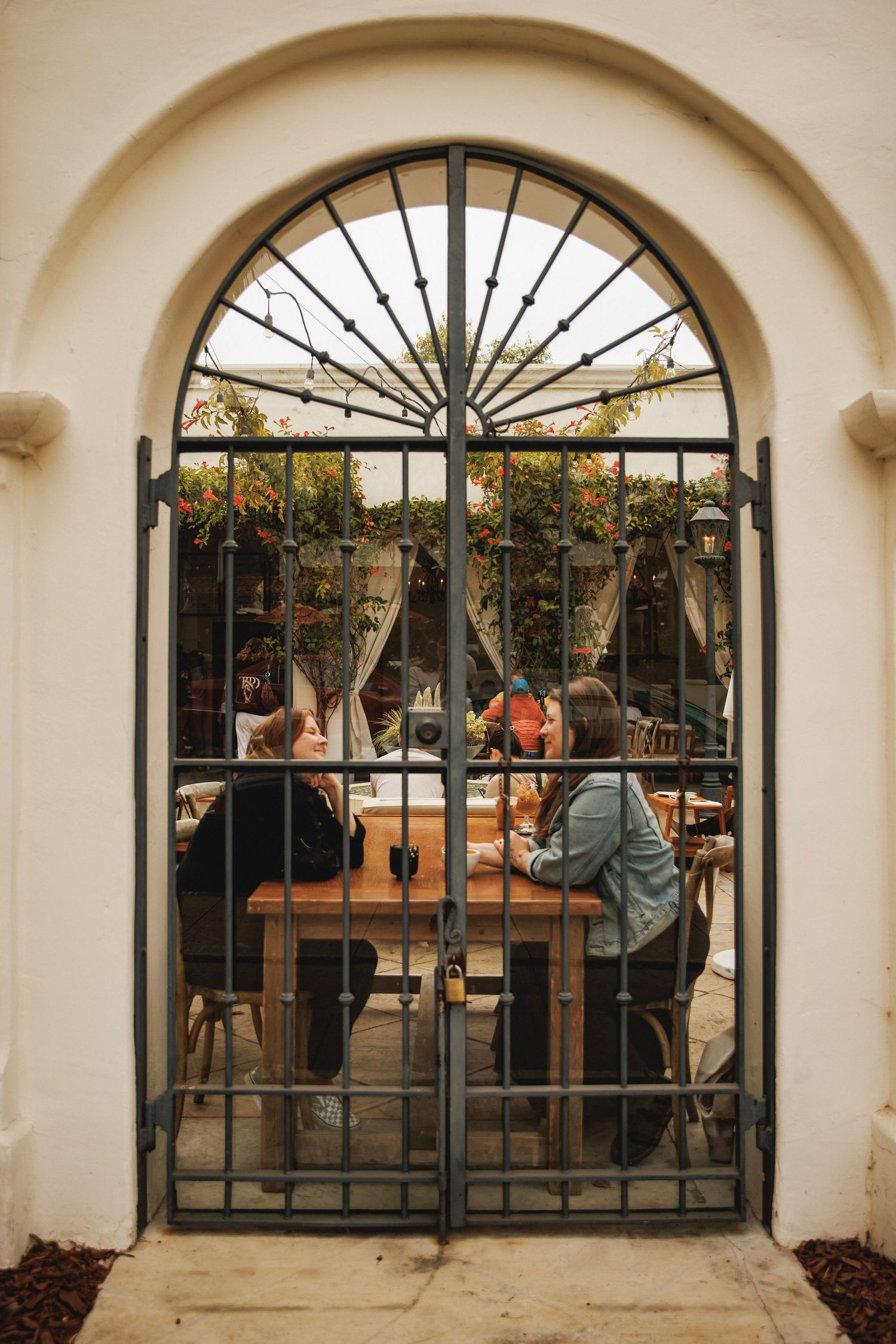 Two women dining at an outdoor patio restaurant viewed through an ornate arched iron gate.