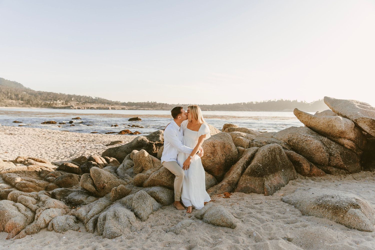 Couple kissing while seated on coastal rocks during a beach engagement session.