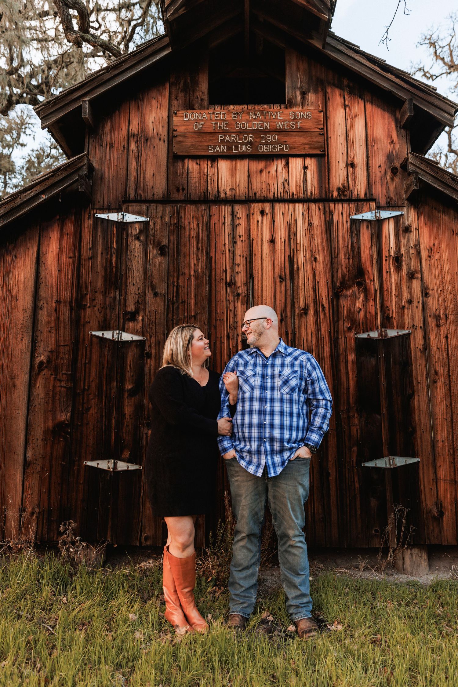 Couple smiling at each other in front of a rustic red barn, woman in orange boots.