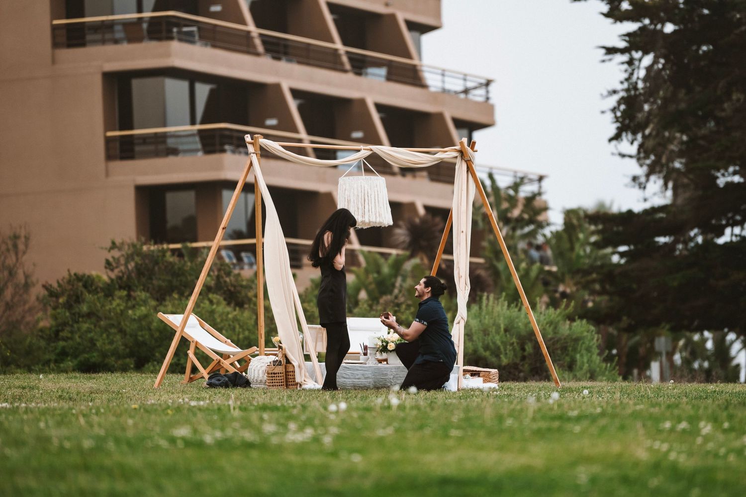 Couple sharing a romantic outdoor proposal moment under a wooden arch on a green lawn with a city building backdrop.
