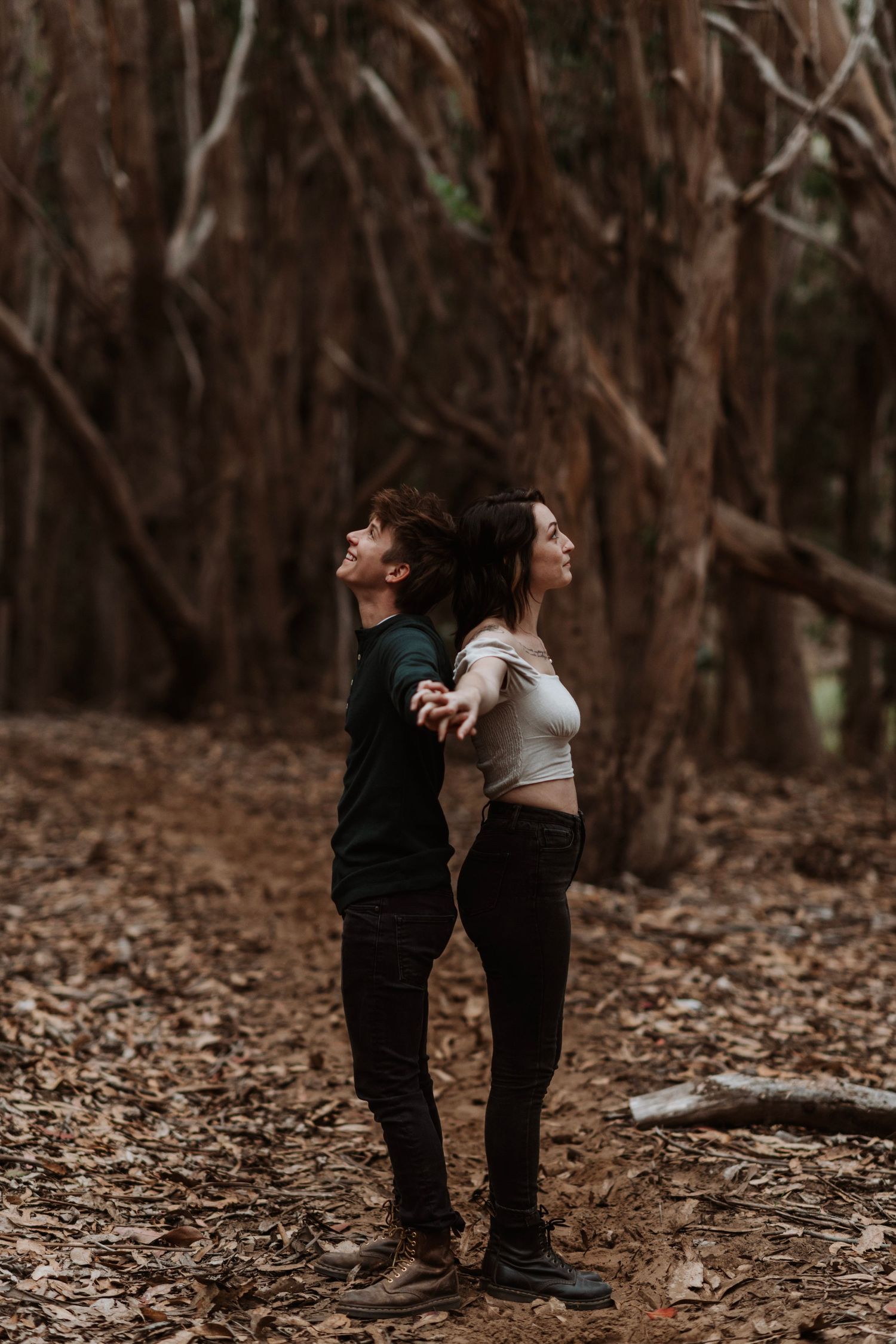 A couple stands back to back in a moody forest, surrounded by bare trees and autumn leaves.