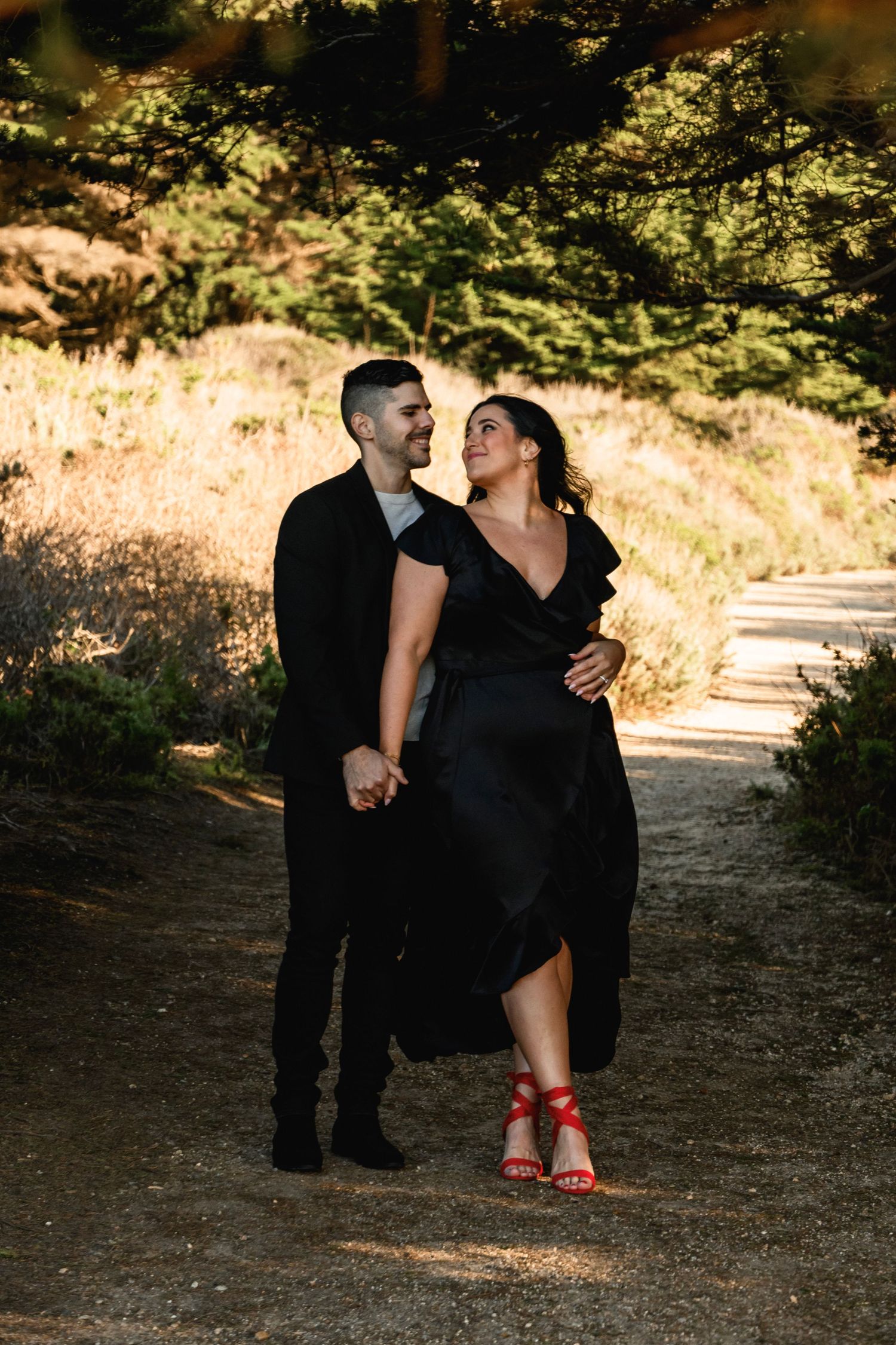 Couple dressed in black holding hands and gazing at each other on a sunlit forest path.