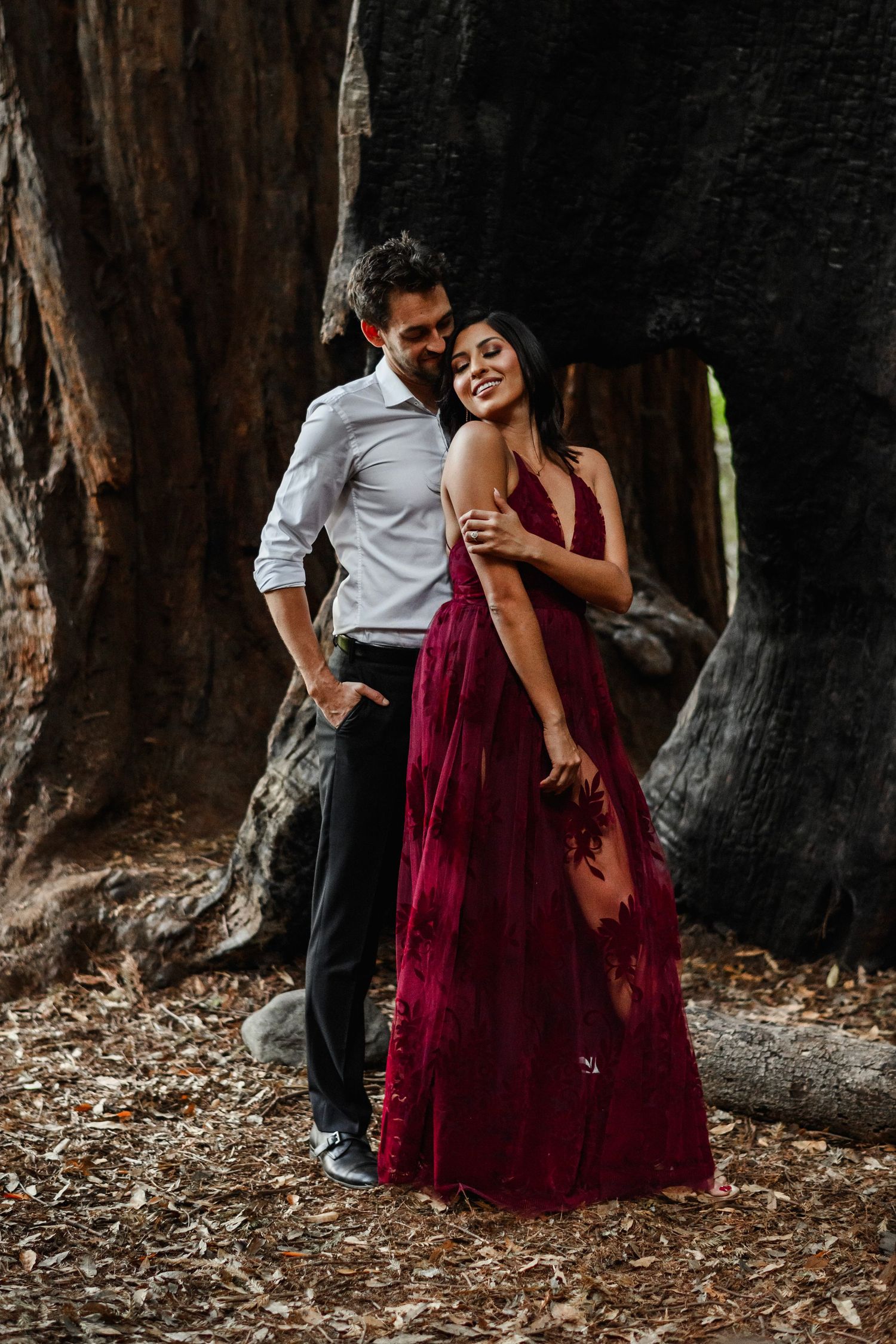 A couple embraces near a massive redwood tree, the woman wearing an elegant burgundy gown in a forest setting.