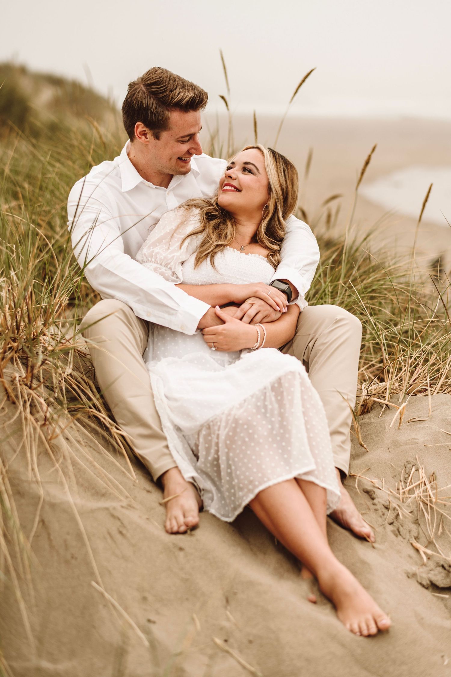 Happy couple sitting on sandy beach dunes embracing and smiling at each other during engagement photos.