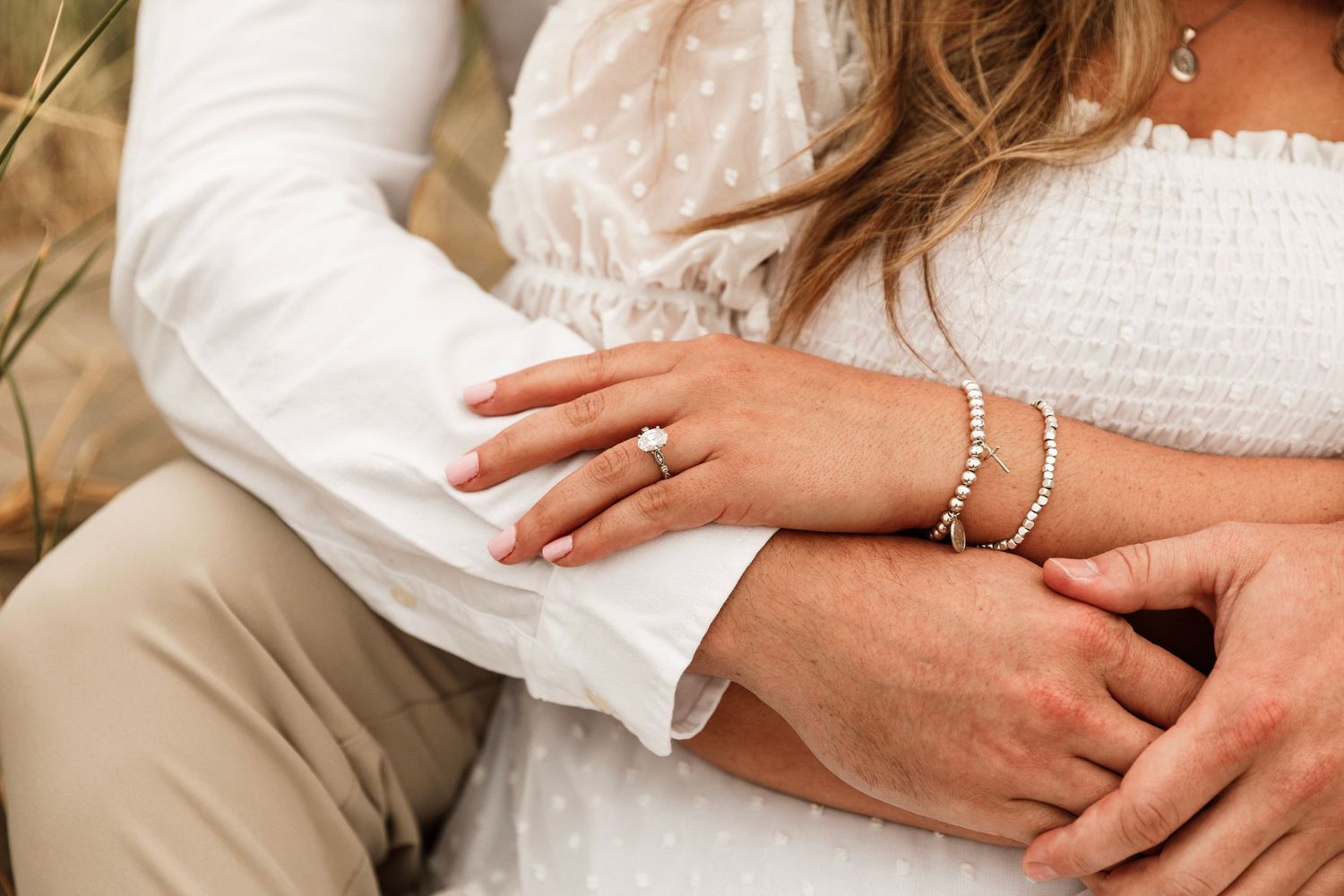 Close-up of couple holding hands showing engagement ring and pearl bracelets during beach engagement session.