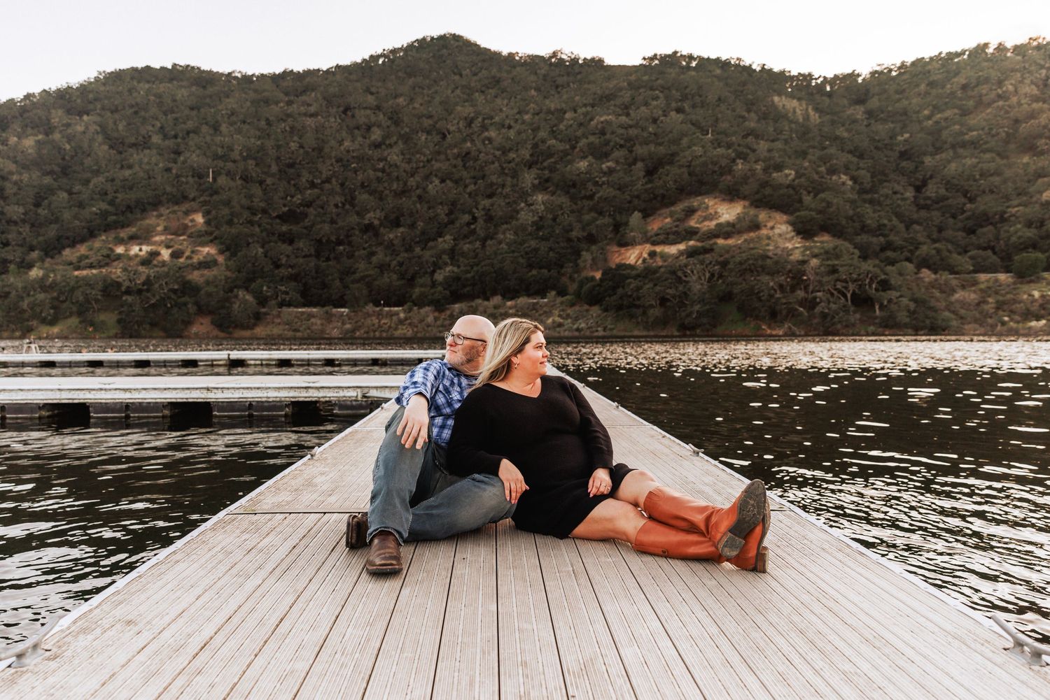 Couple sitting together on a lake dock at dusk with forested hills in the background.