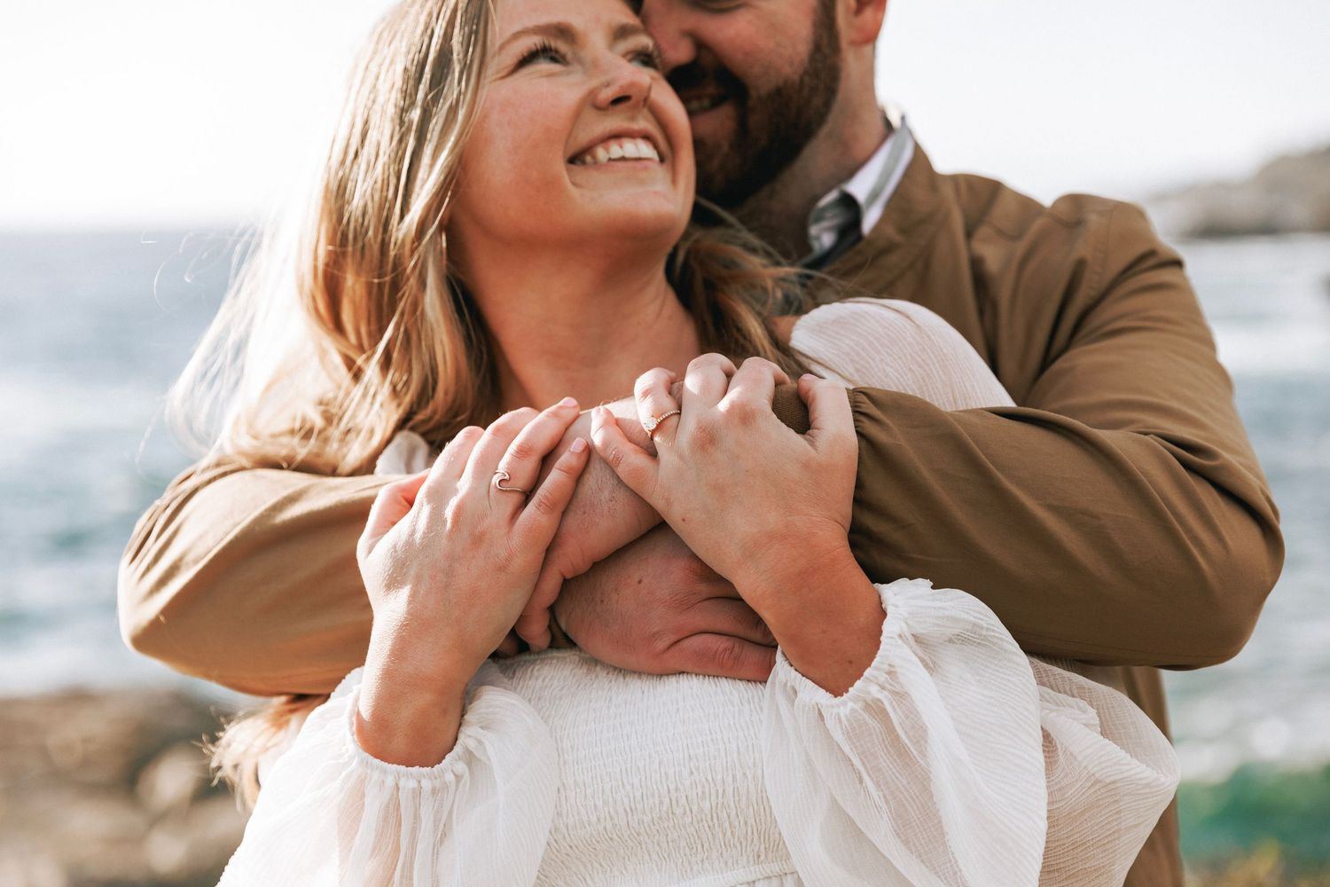 Man tenderly embracing woman from behind near rocky coastal scenery in warm sunlight.
