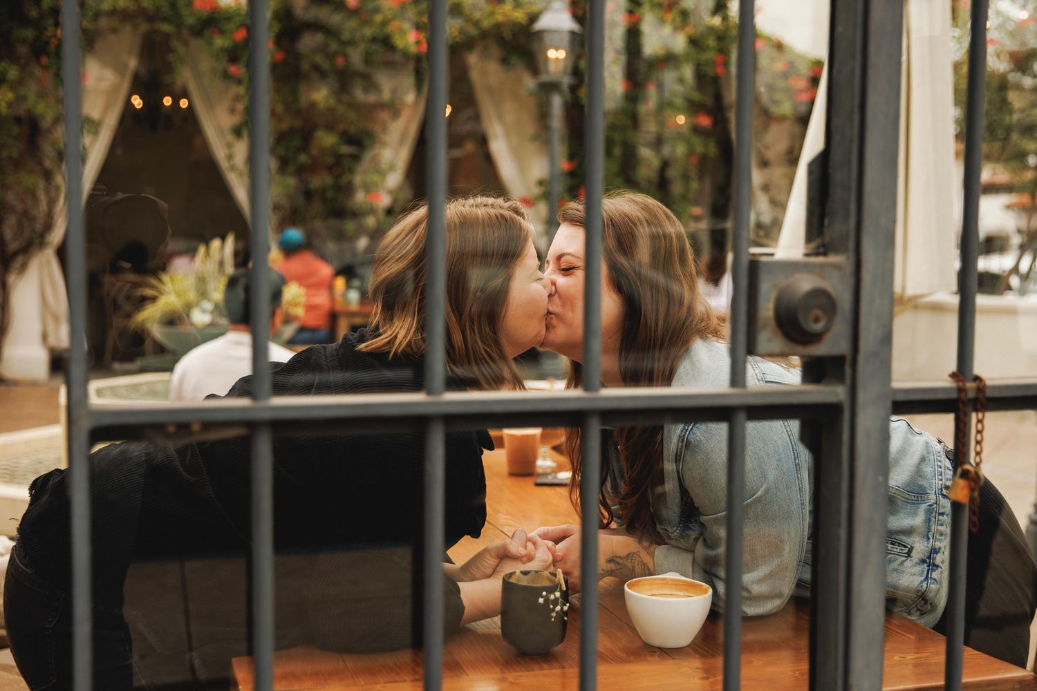Couple sharing a kiss at outdoor cafe table, viewed through decorative iron fence bars.