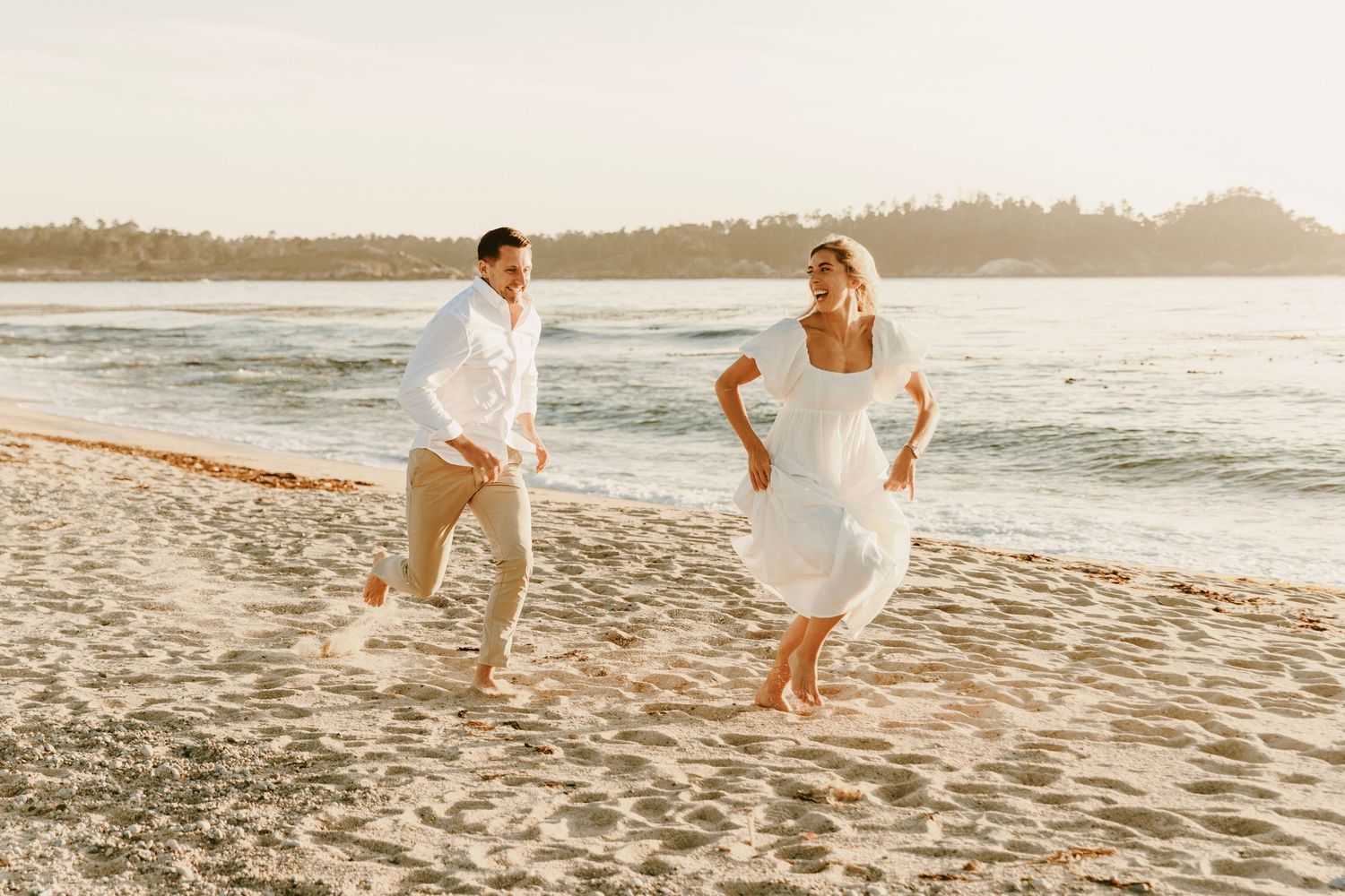 Couple in white running playfully along a sunny sandy beach at golden hour.