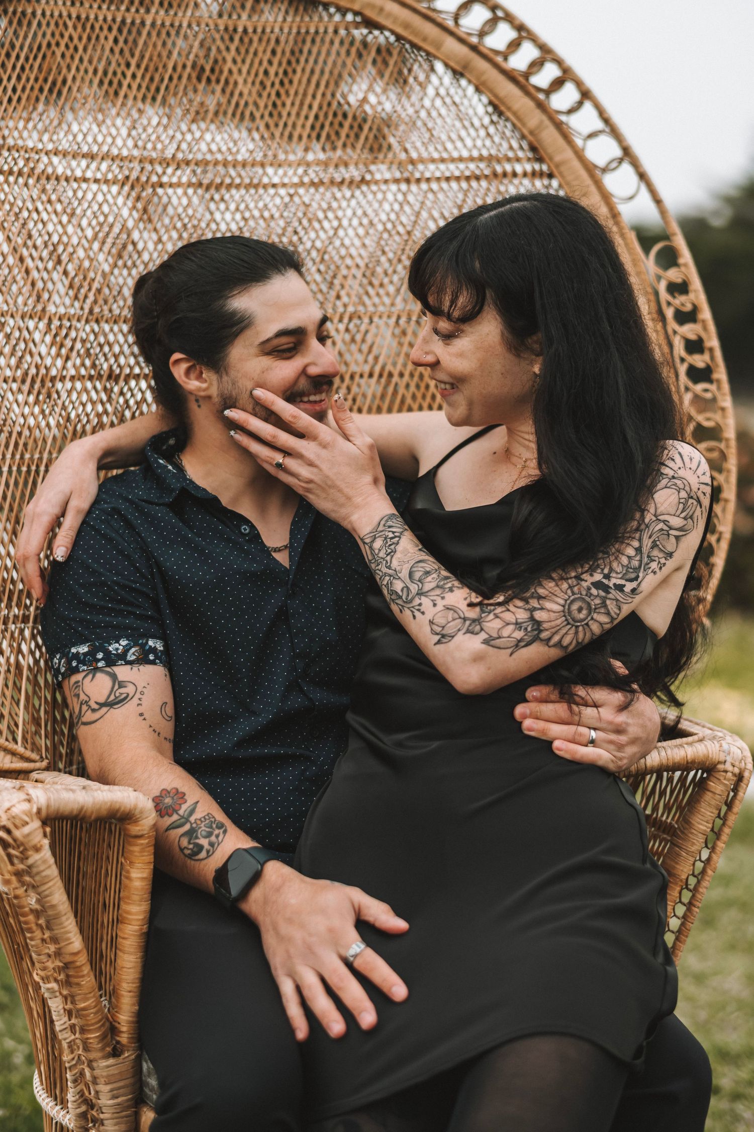 A tattooed couple shares a playful intimate moment seated in a large wicker peacock chair outdoors.