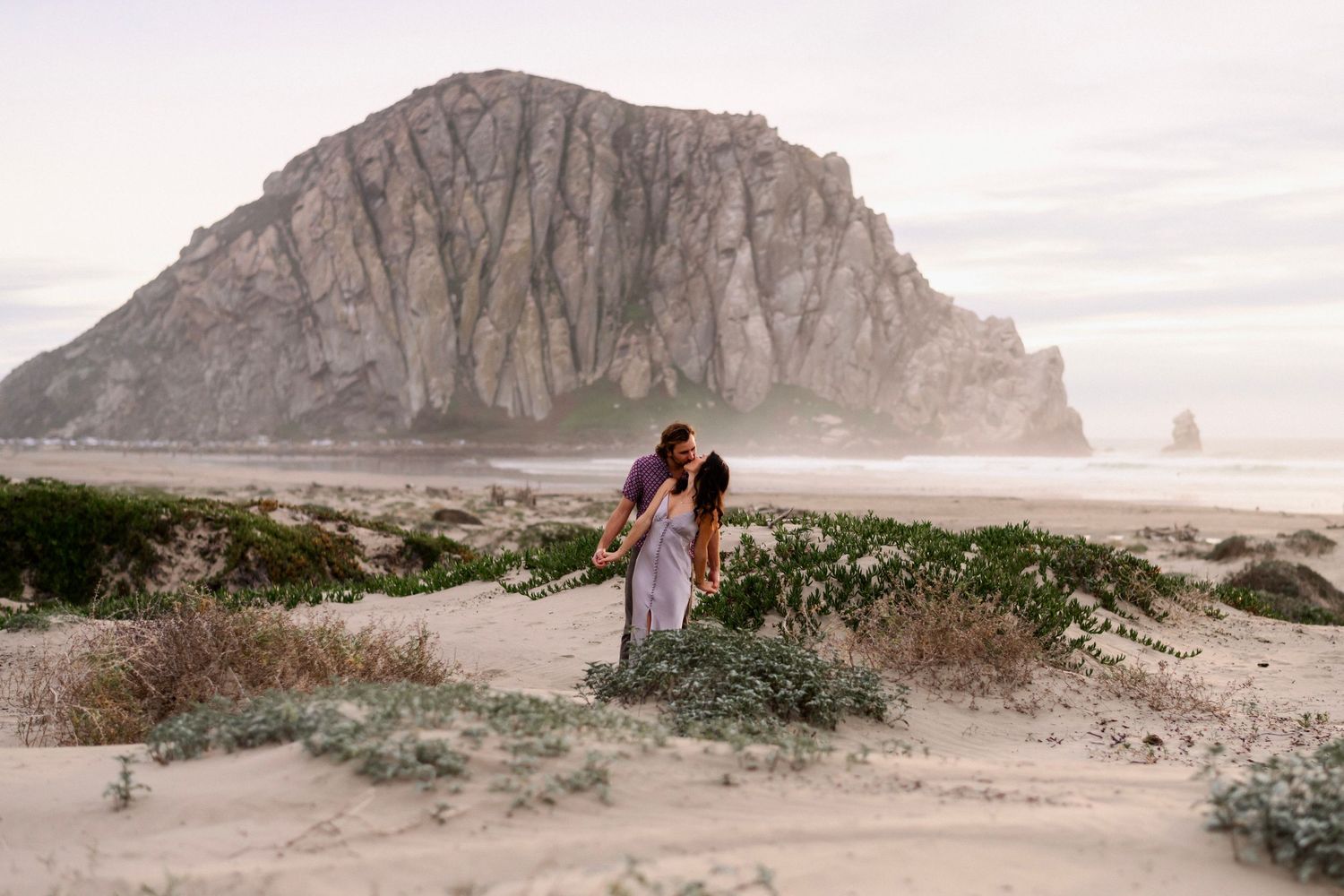 Couple embracing on sandy beach with iconic Morro Rock in background.