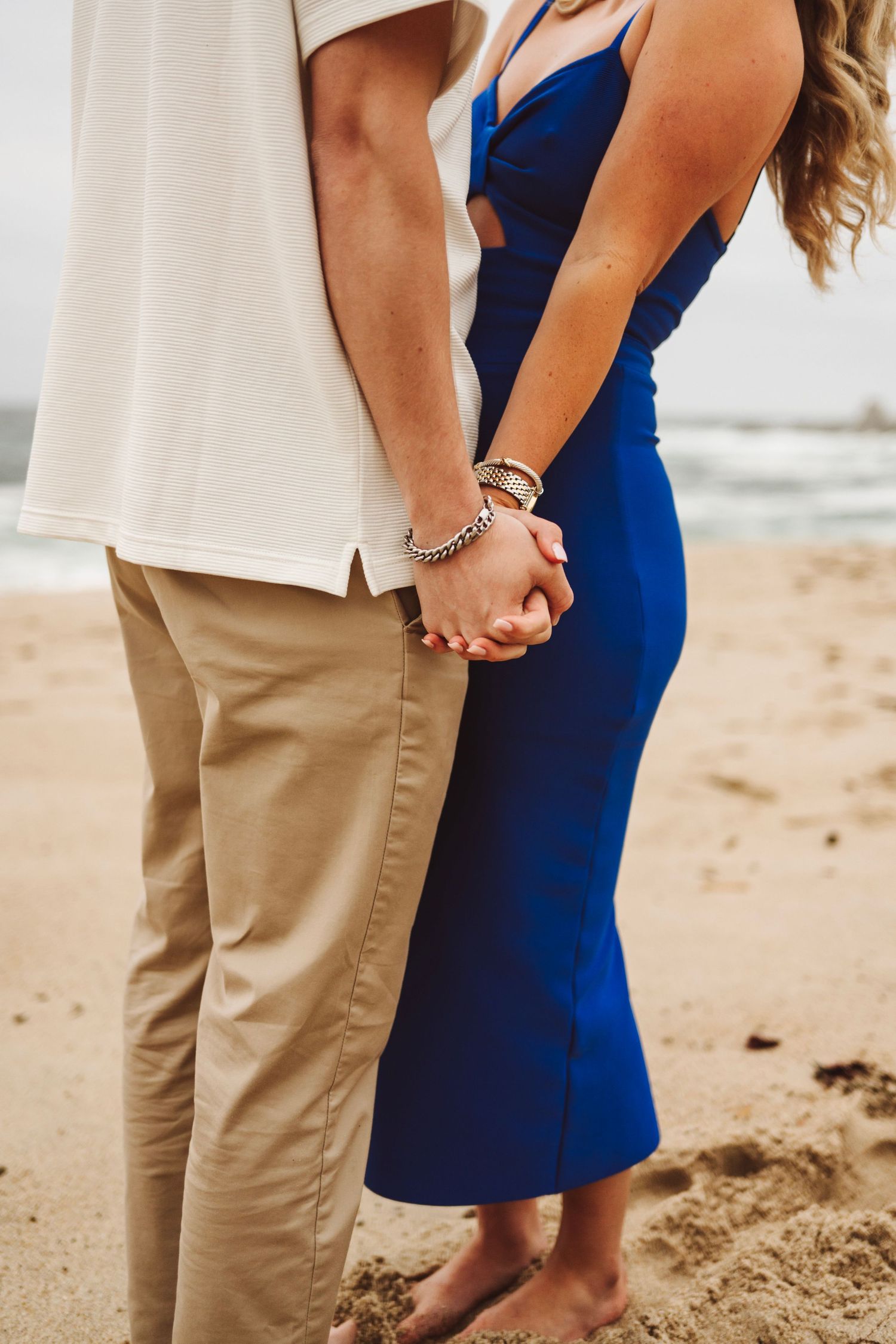 Couple holding hands on sandy beach, woman in blue dress and man in khaki pants with white shirt.