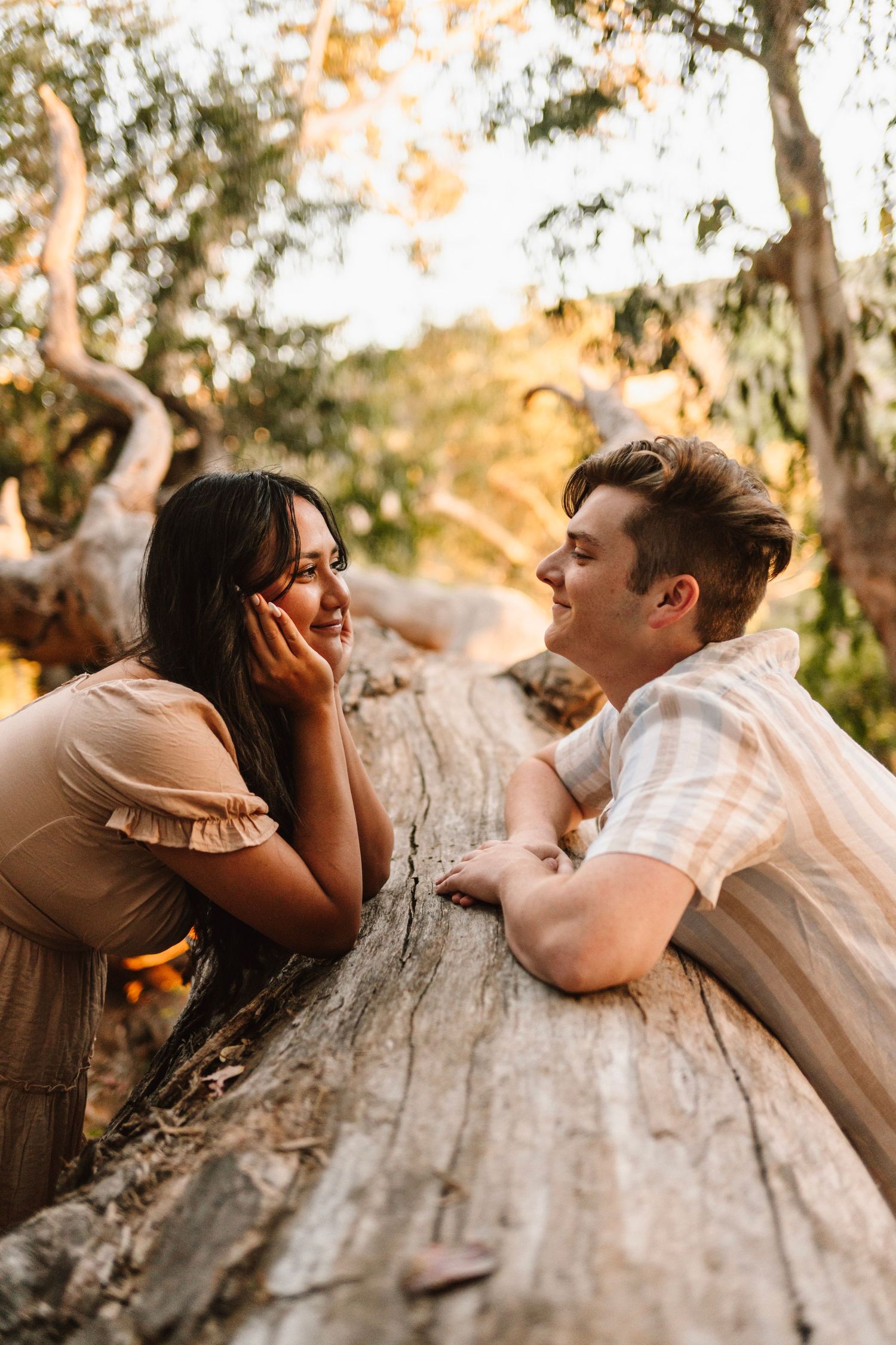 Couple leaning on a fallen log in a golden forest, sharing a tender moment at sunset.