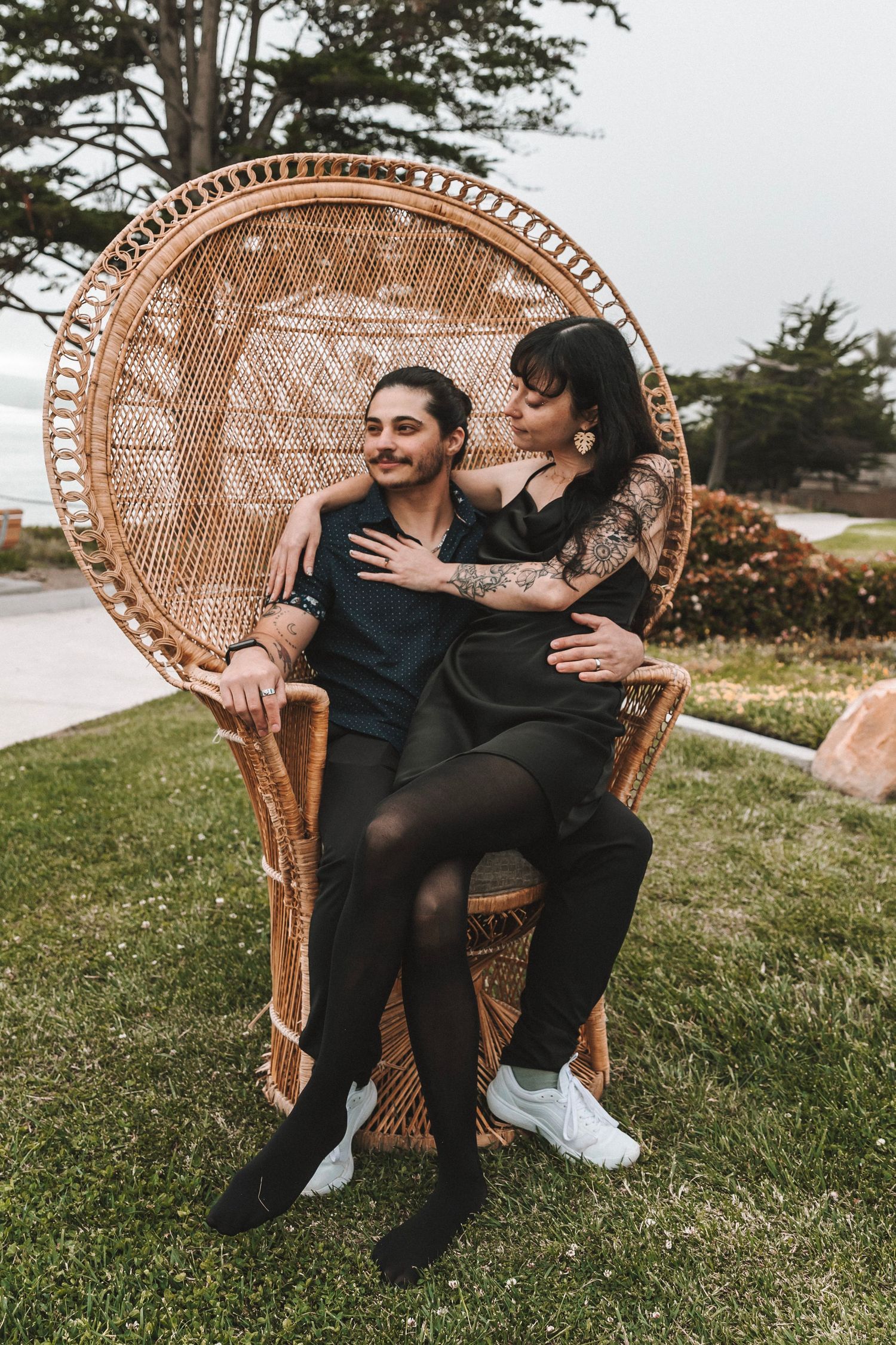 A smiling couple poses together in a large decorative wicker peacock chair on a green lawn.