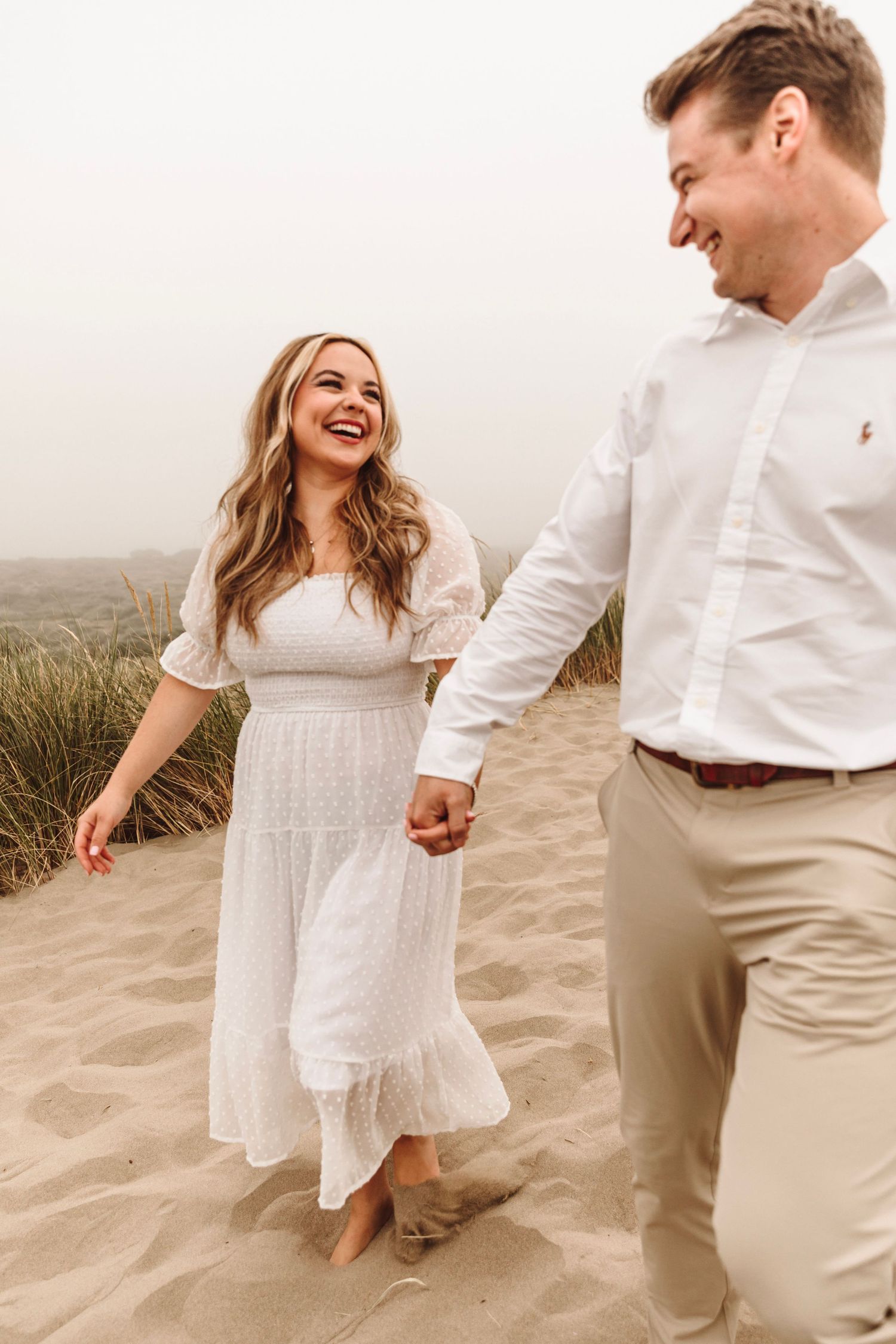 Joyful couple holding hands and laughing while walking barefoot on sandy beach dunes together.