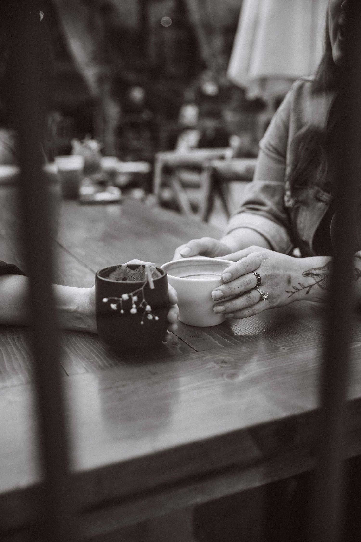 Black and white close-up of two people holding coffee cups at a wooden cafe table.
