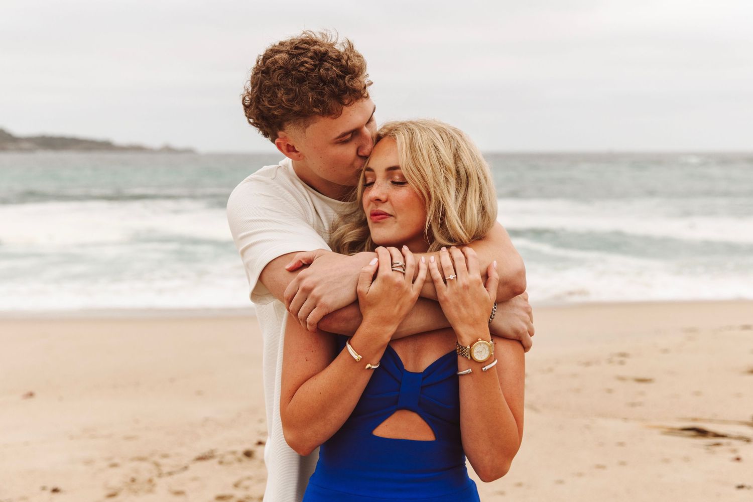 Young couple embracing on a beach with ocean waves, woman wearing a blue swimsuit.
