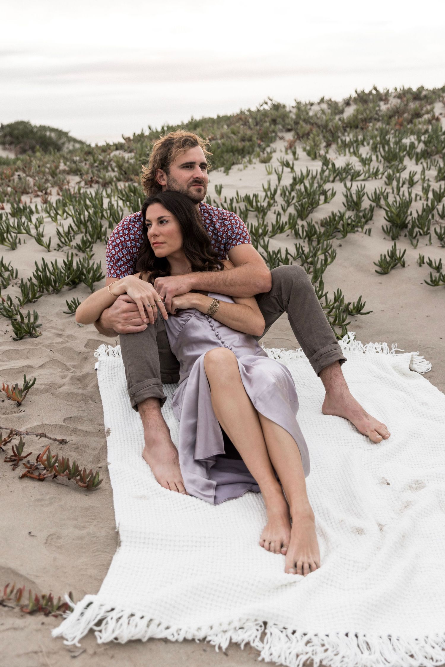Couple sitting on white blanket on sand dunes surrounded by coastal plants.