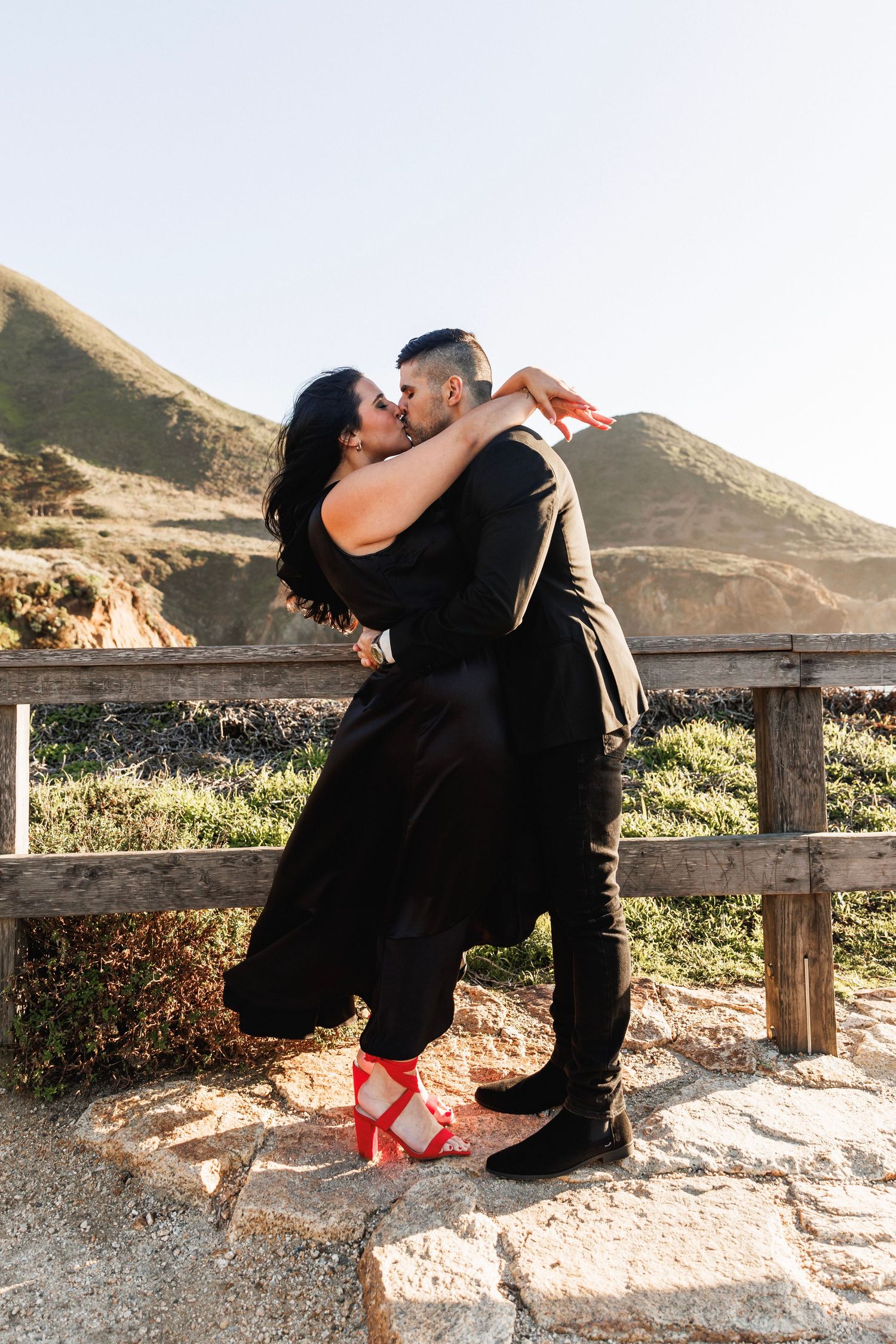 Couple in matching black outfits sharing a passionate kiss with scenic mountain backdrop behind them.