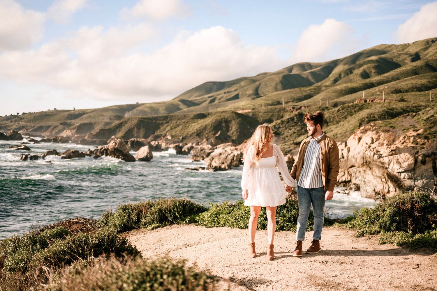 Couple walking hand in hand along a scenic coastal path with green cliffs and ocean.