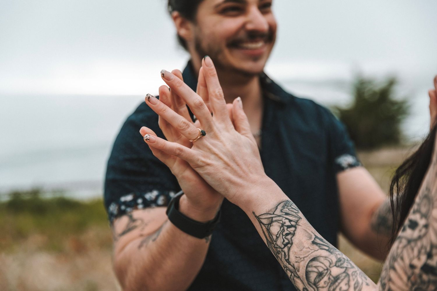 A close-up of a tattooed couple holding hands and displaying their engagement rings outdoors.