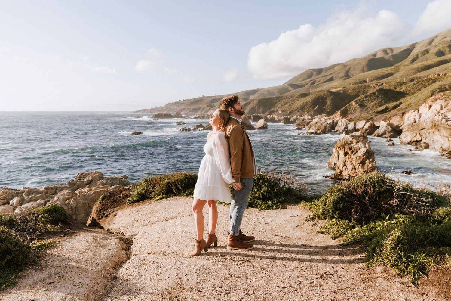 Couple holding hands on coastal trail overlooking rocky Pacific Ocean shoreline on sunny day.