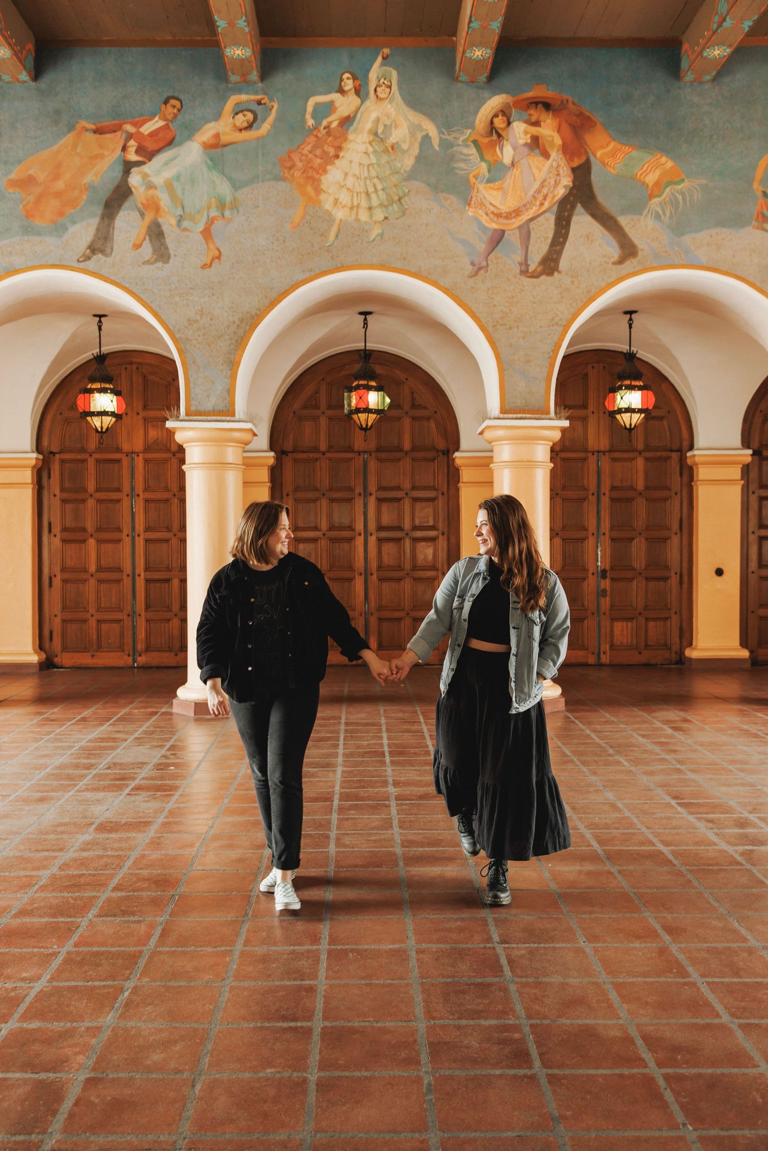 Two women holding hands and walking through an elegant hall with colorful murals and arches.