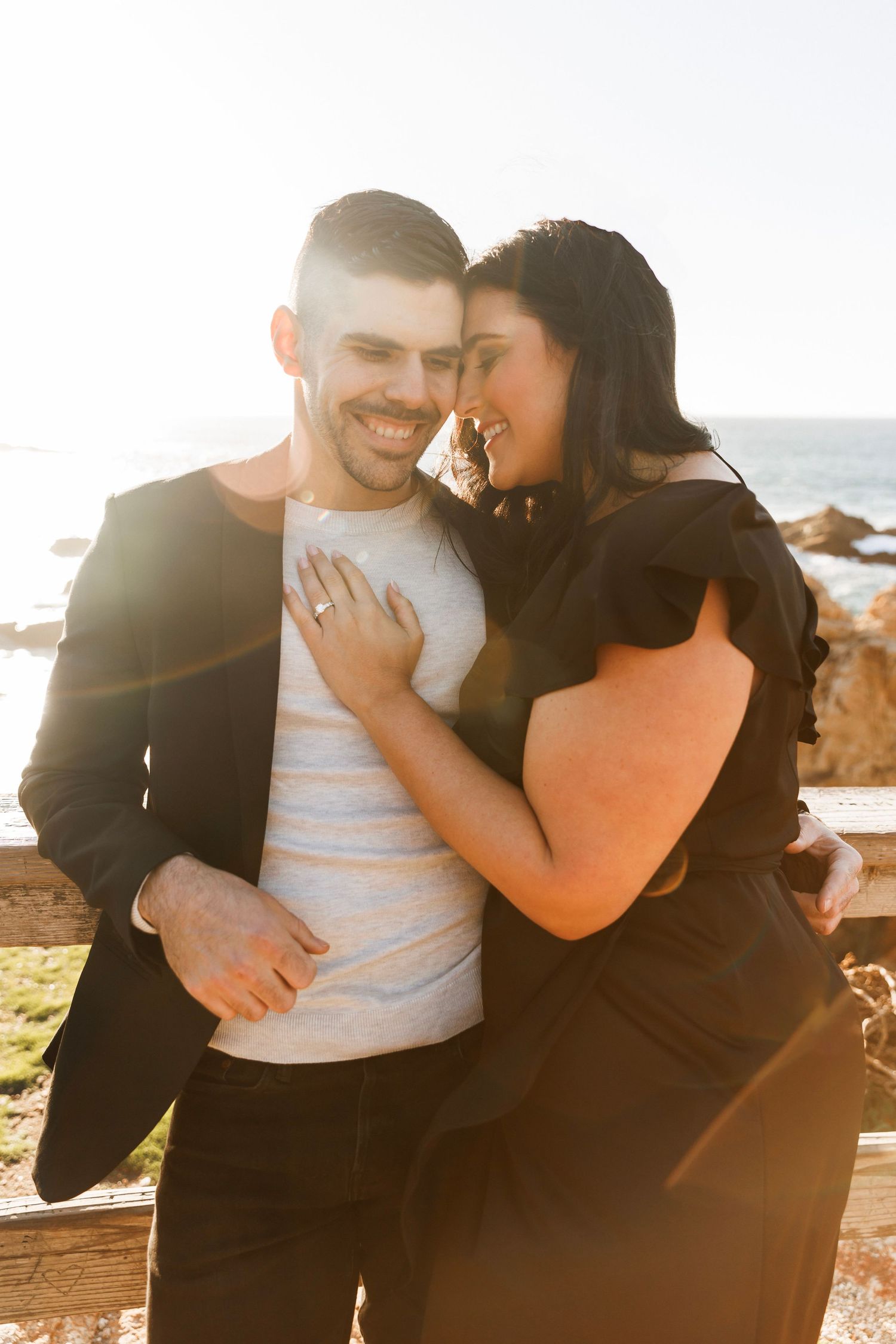 Smiling couple embracing in golden sunset light near ocean overlook during engagement photo session.