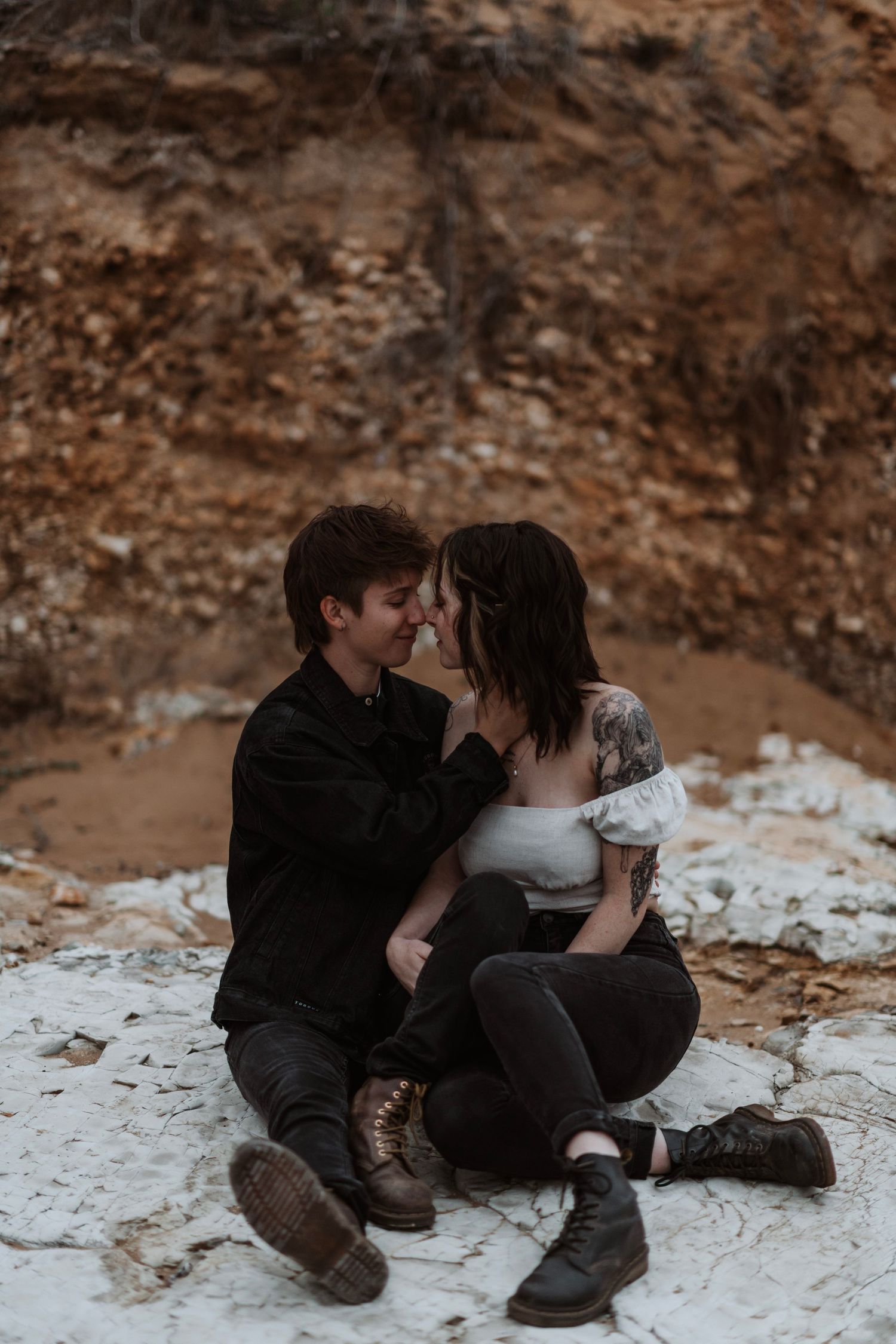 Couple sitting close together on white rocks with sandy cliffs behind them.