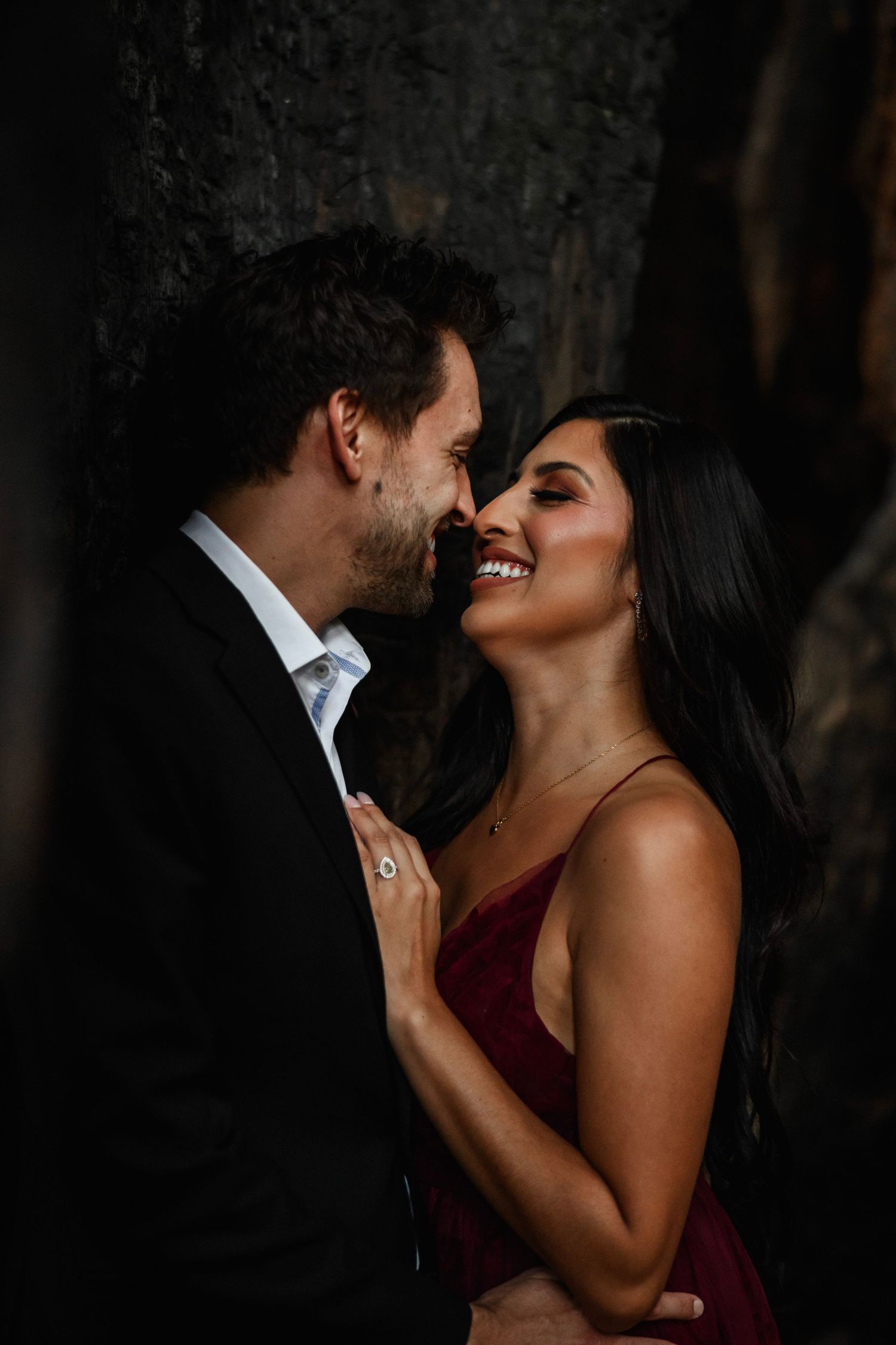 Romantic couple nearly kissing in dramatic dark setting, woman in red dress smiling at bearded man in black suit.