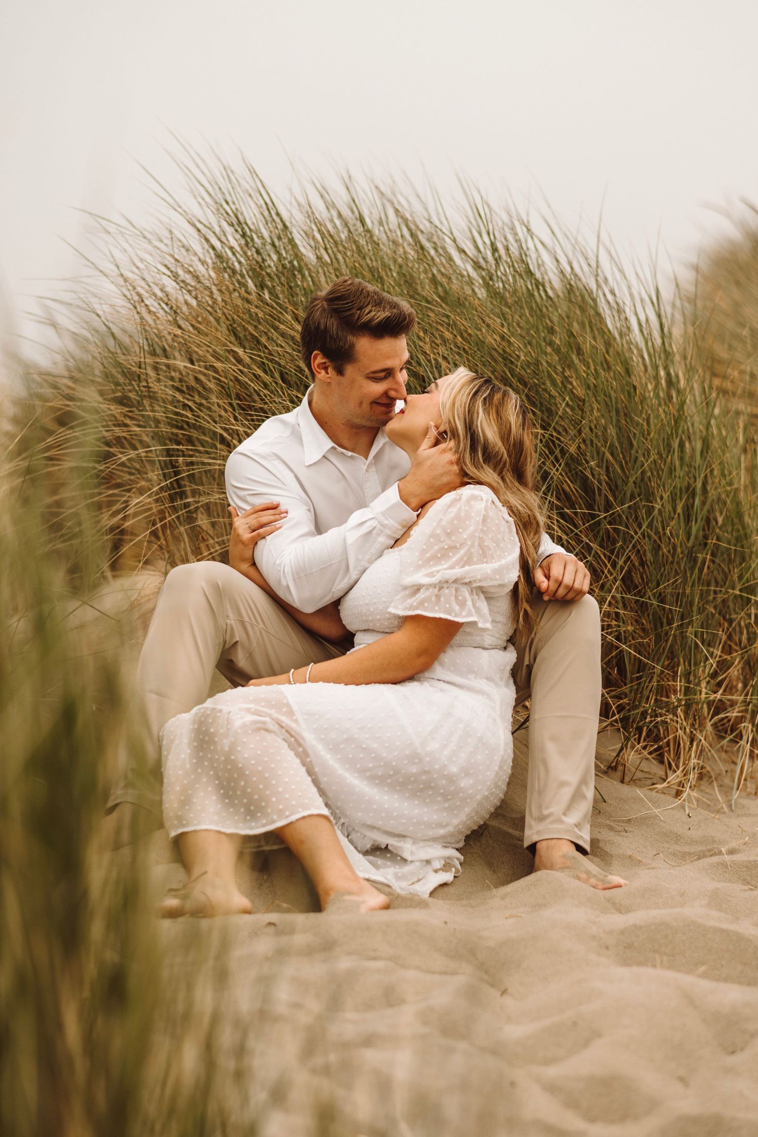 Romantic couple embracing and kissing among tall beach grass sand dunes on overcast day.