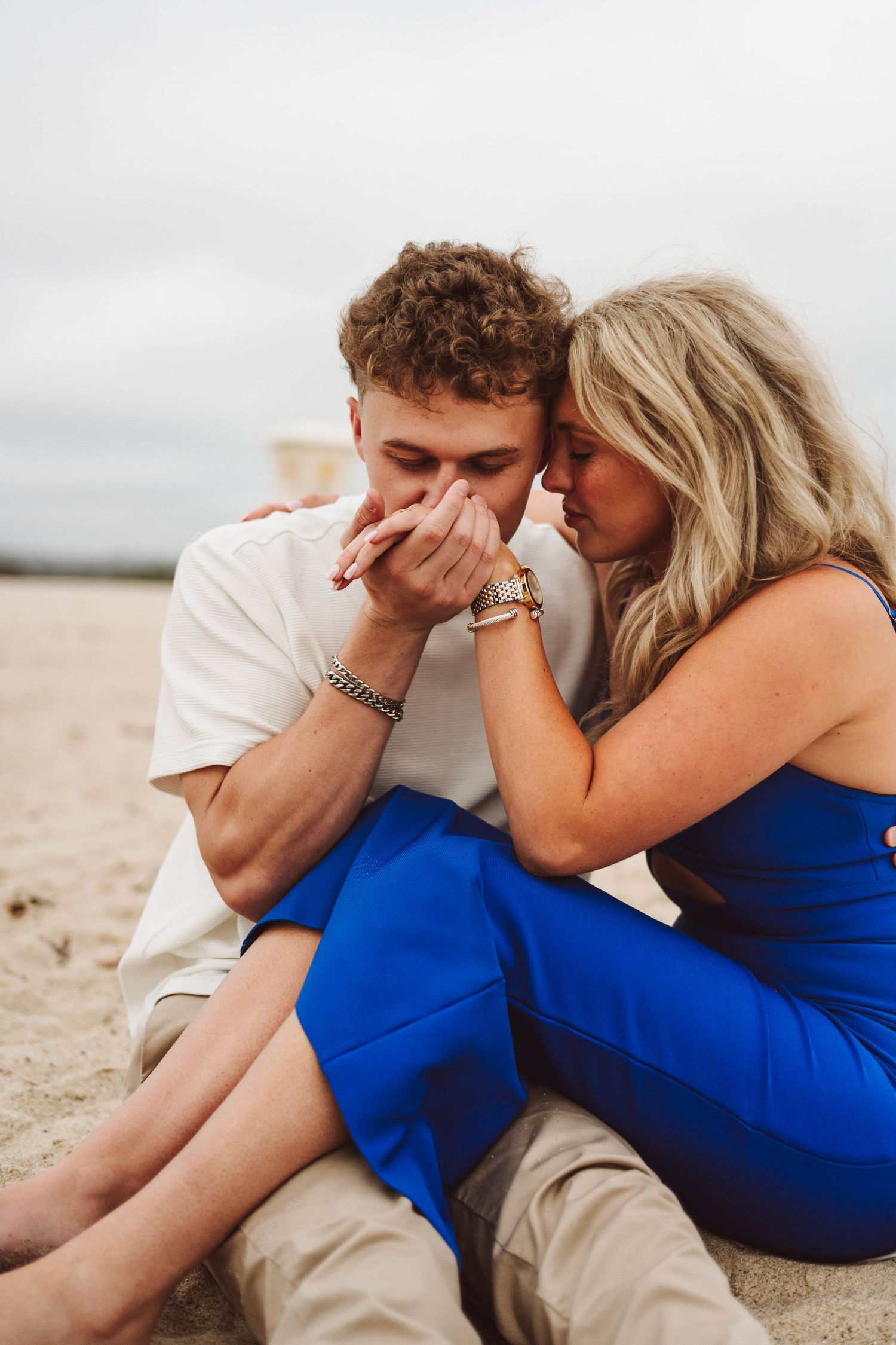 Romantic couple sitting on beach sand, man kissing woman's hands while she leans close in blue dress.