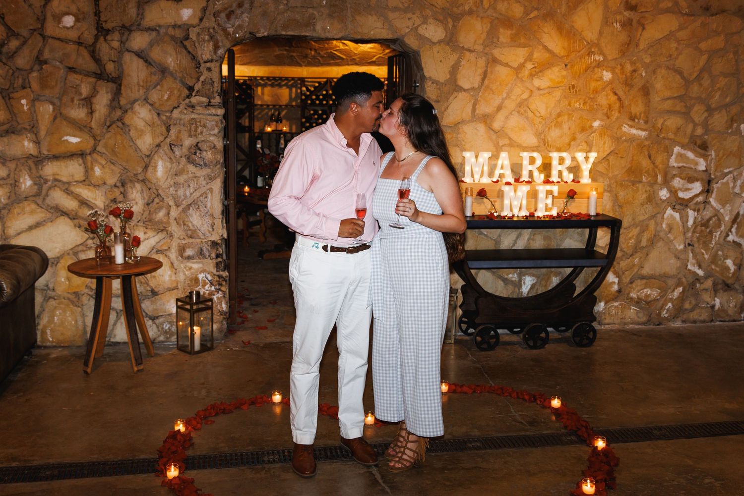 Couple embraces near glowing Marry Me marquee sign surrounded by candles in romantic stone venue.