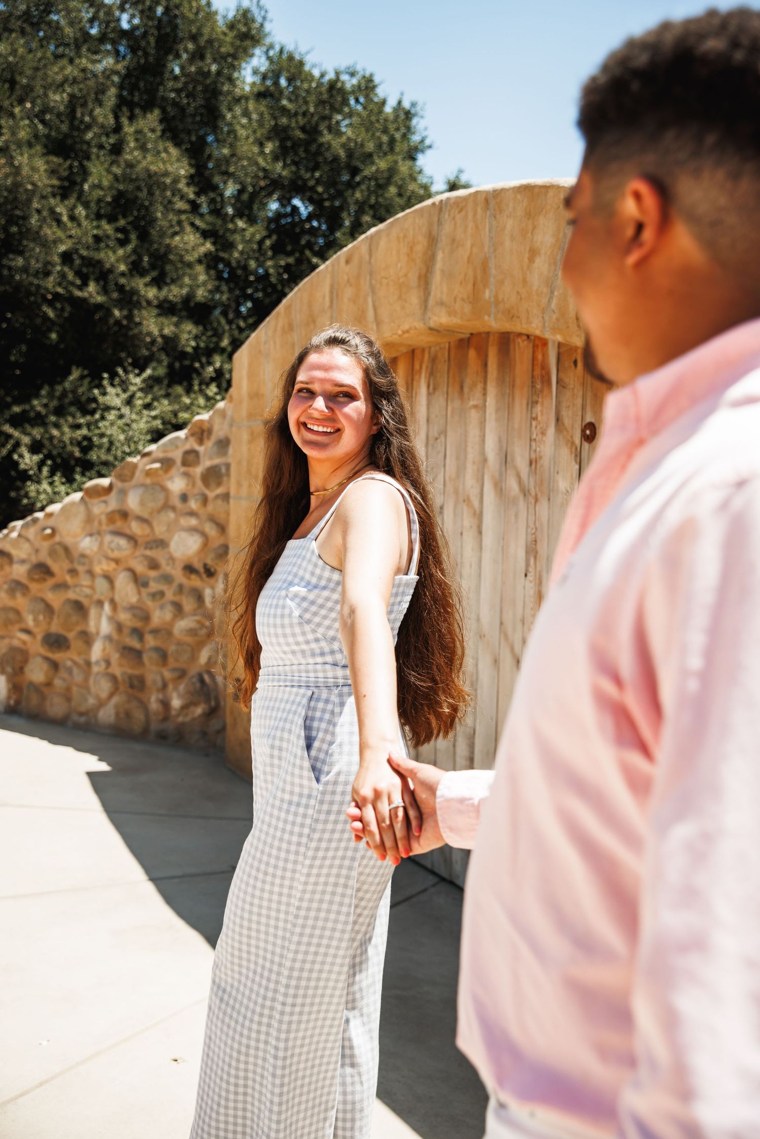 Smiling woman in blue sundress holds hands with partner outdoors near stone wall on sunny day.