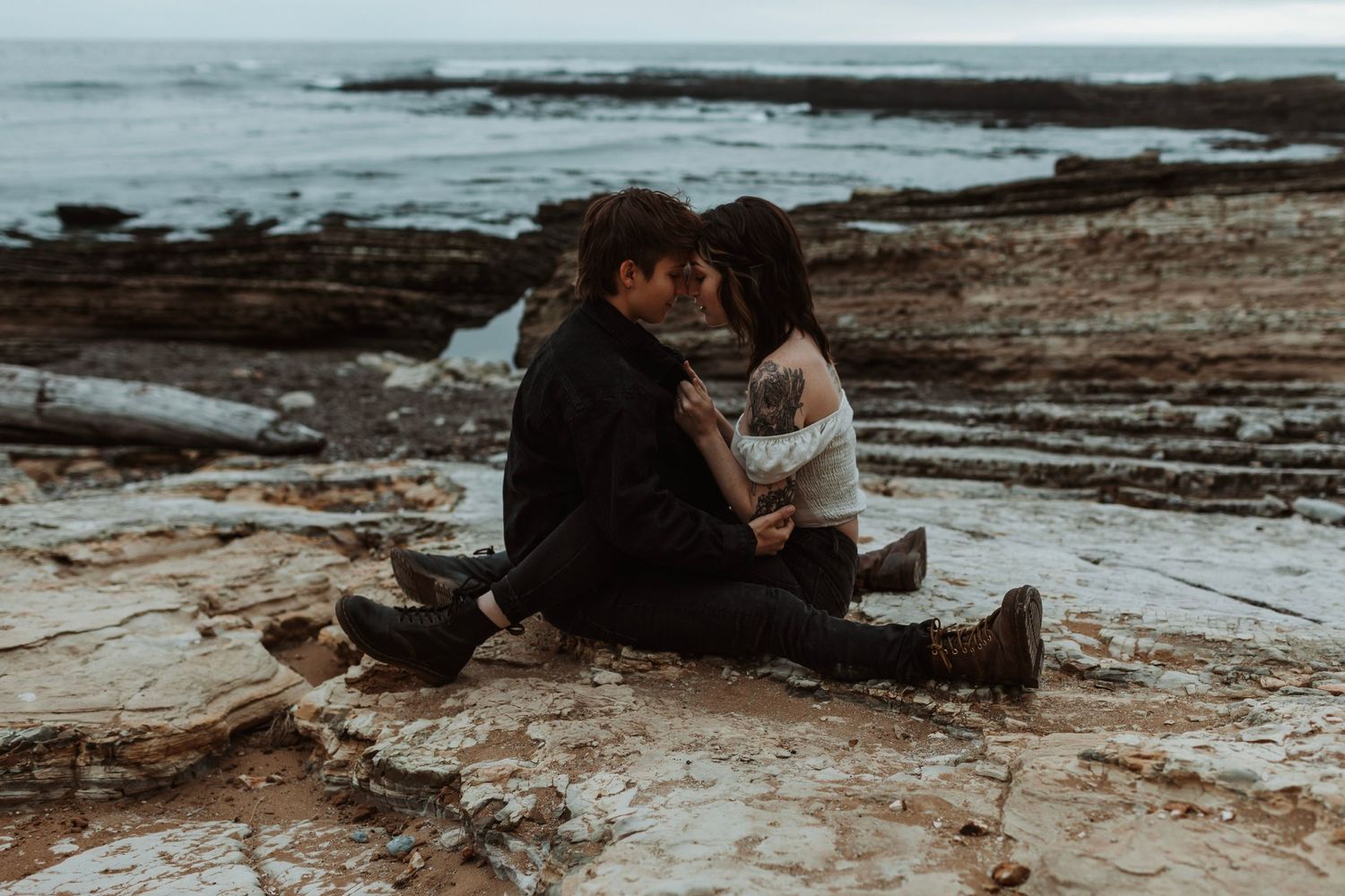 Couple sitting intimately on rocky beach shore, leaning foreheads together in a quiet romantic moment.
