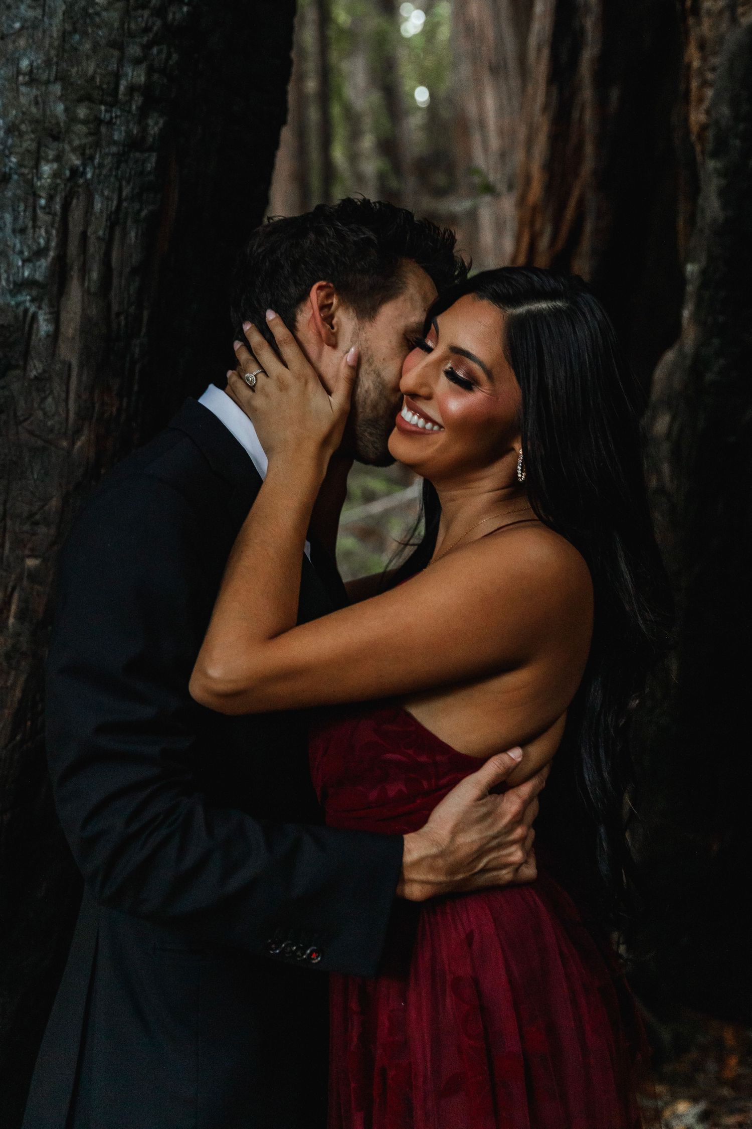 A smiling couple embraces passionately against a dark redwood tree, the woman in a stunning red lace gown.