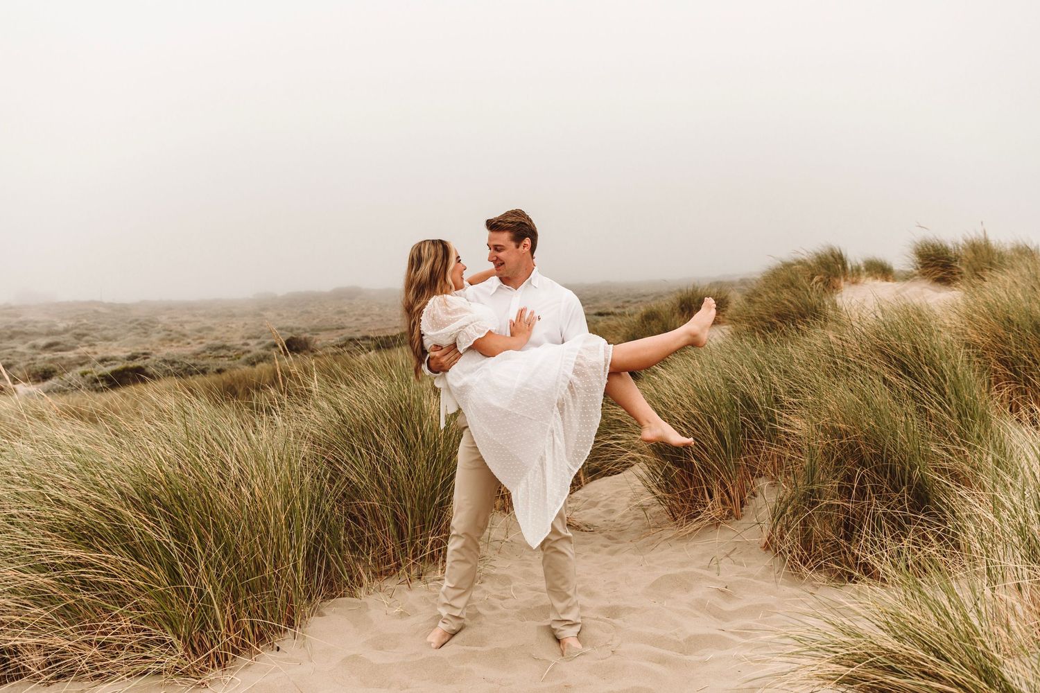 Man lifting woman in white dress among beach grass dunes during romantic engagement photo session.