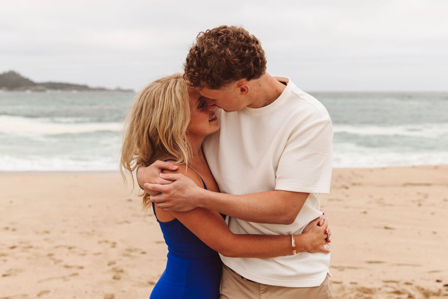 Couple embracing and kissing on a sandy beach with ocean waves in the background.