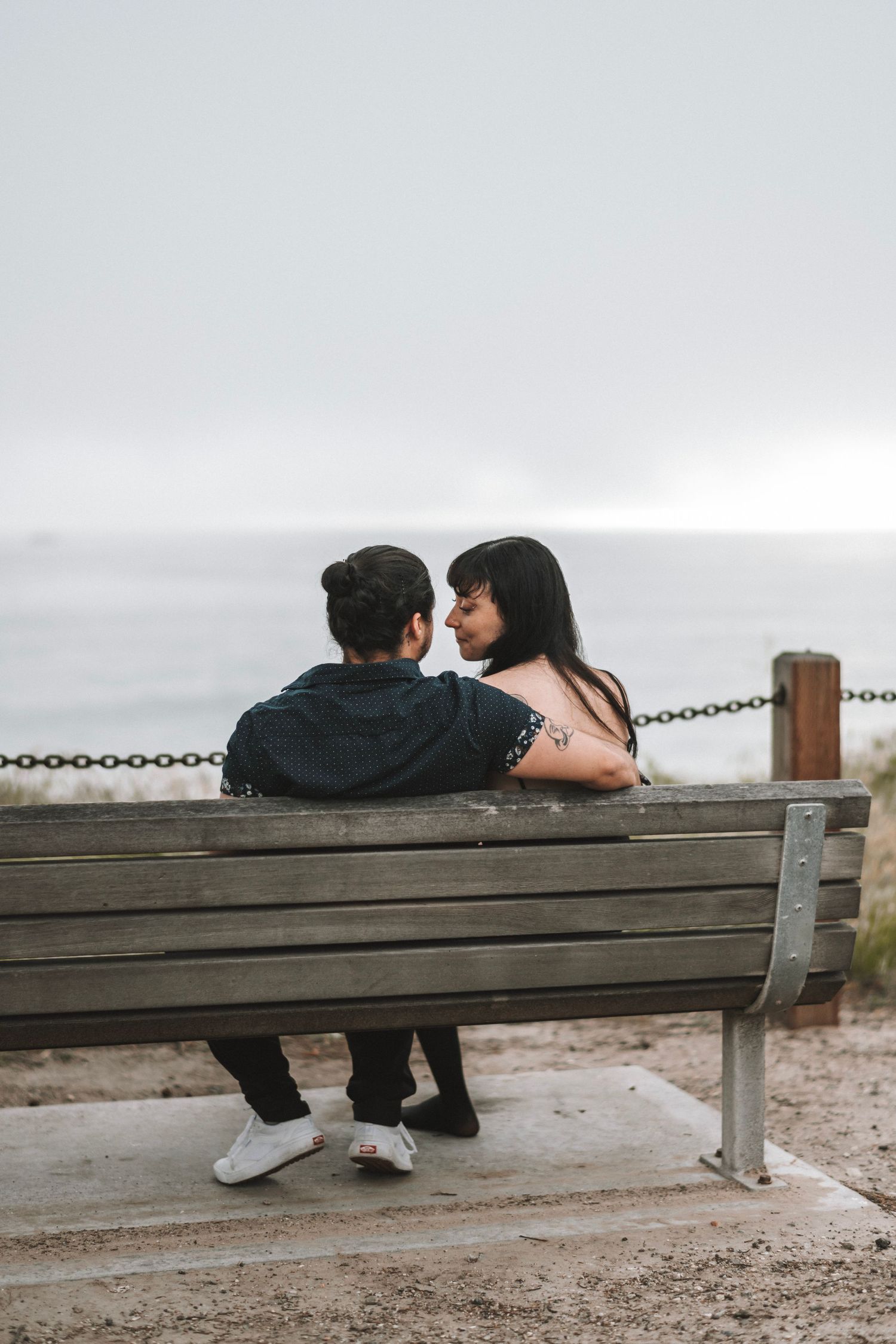 A couple sits closely on a wooden bench facing the ocean, sharing a tender romantic moment.